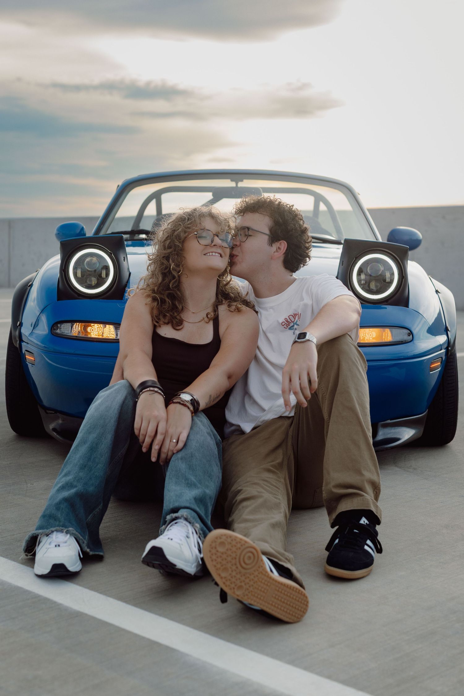 Romantic couple sitting in front of a blue convertible sports car at sunset, sharing a kiss.