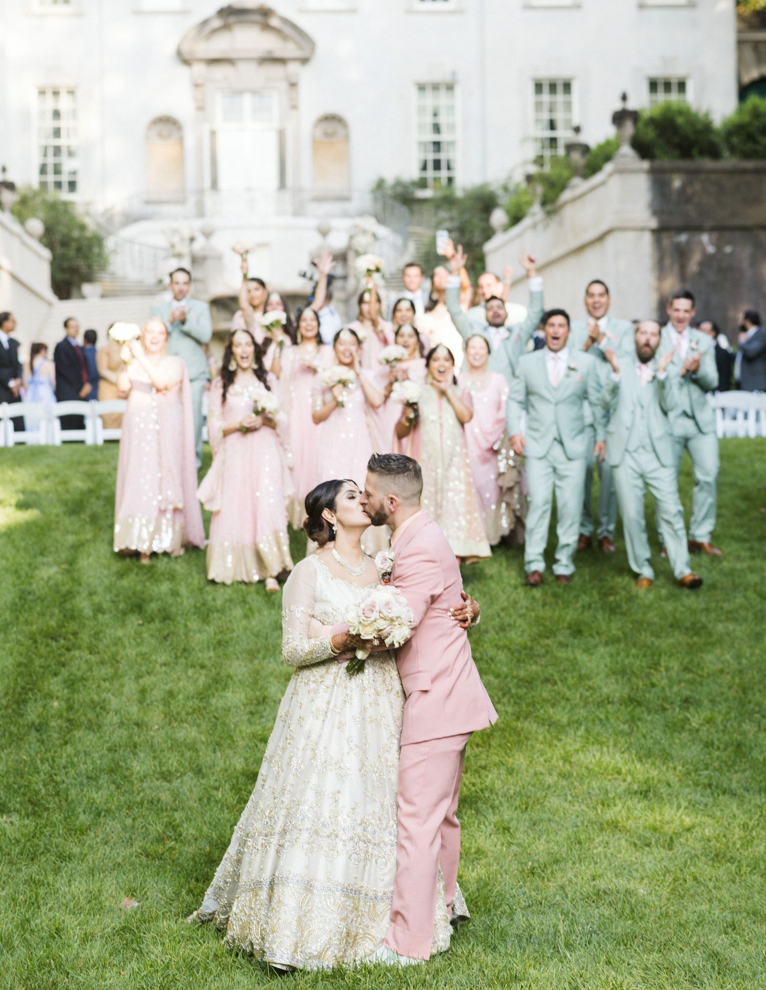 Wedding party in pastel pink and blue attire poses on a grassy lawn in front of an elegant white building.