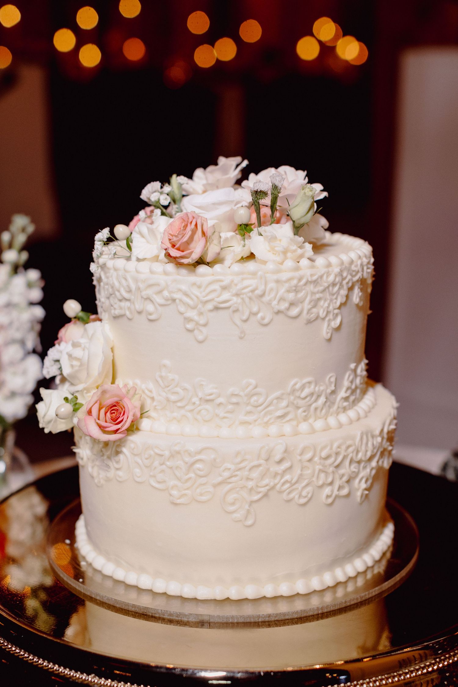 Elegant two-tier white wedding cake decorated with delicate lace piping and fresh pink roses on gold cake stand.