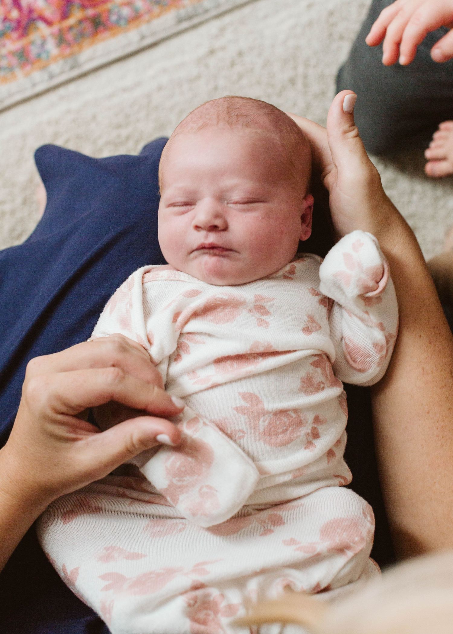 A sleeping newborn baby wearing a white and pink outfit lies peacefully in someone's arms.