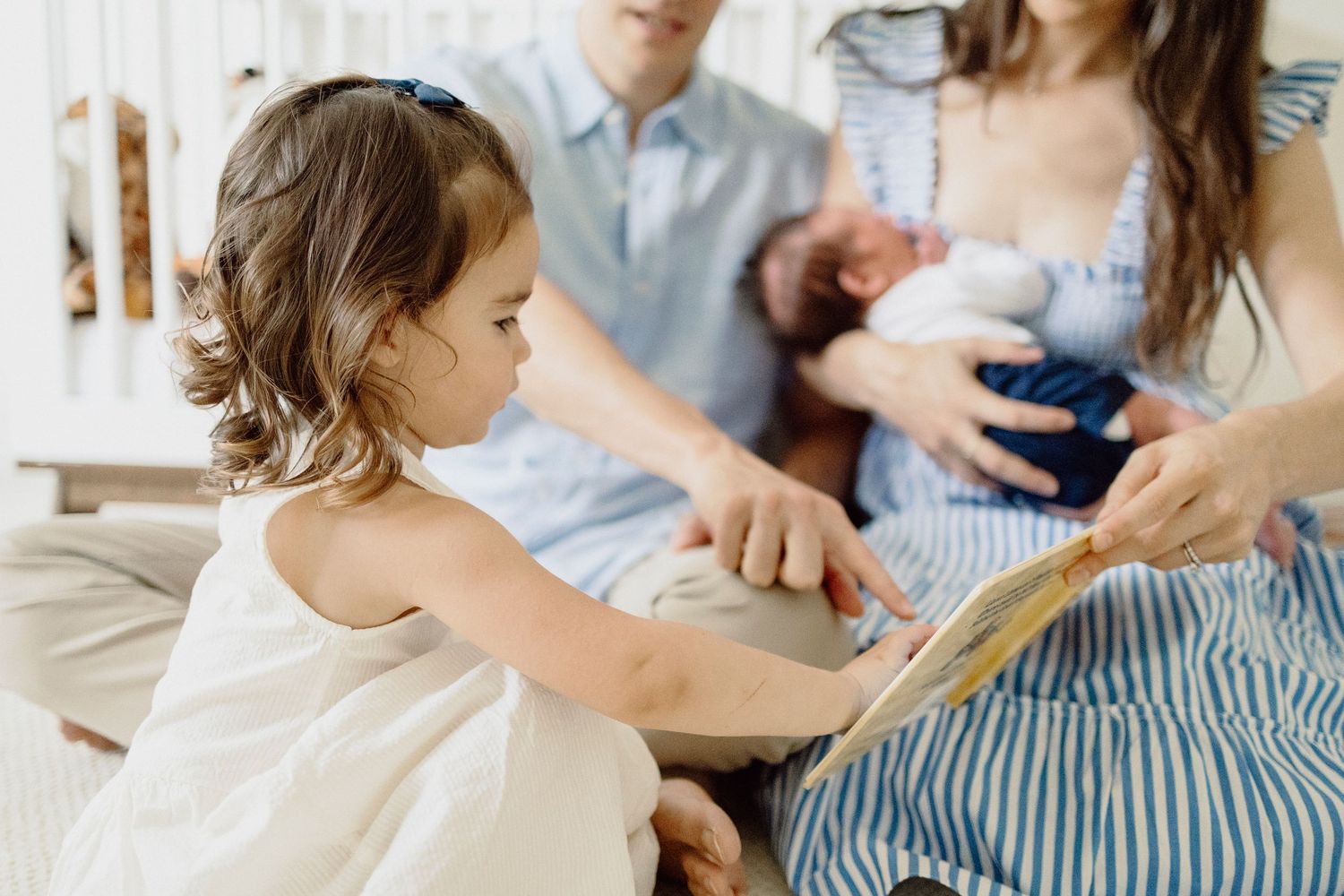 Young child in white dress points to book while family gathers on couch for story time.