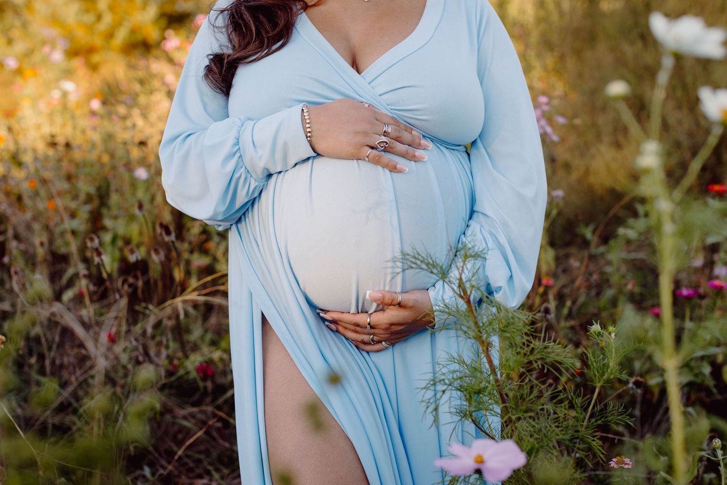 A maternity photoshoot in a light blue dress set against golden autumn foliage in an outdoor garden setting.
