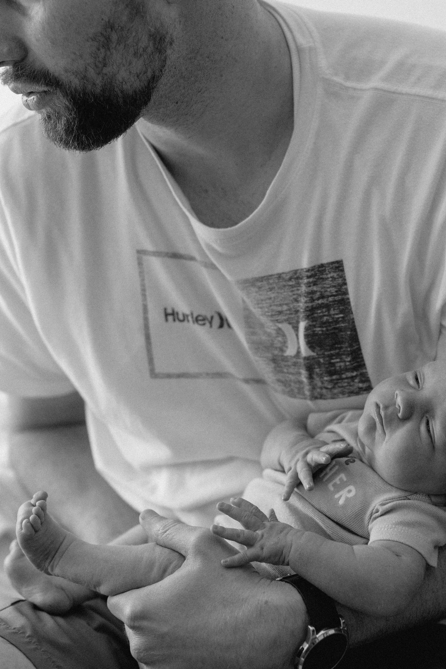 Black and white photo of a bearded man in a Hurley shirt tenderly holding a sleeping newborn baby.