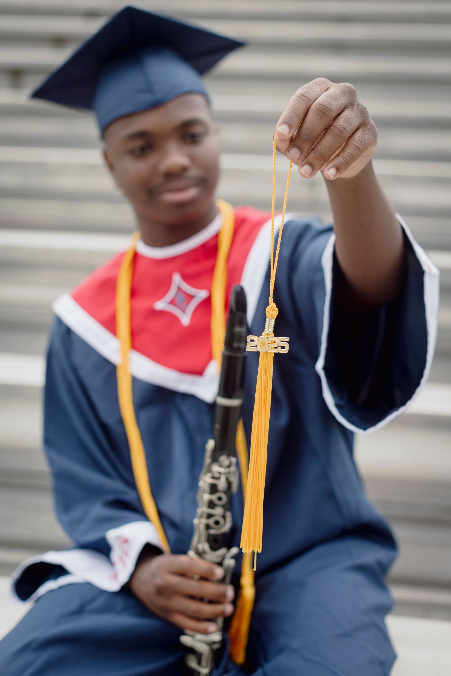 A graduate in blue cap and gown holds a 2025 tassel while clutching a clarinet on bleacher steps.