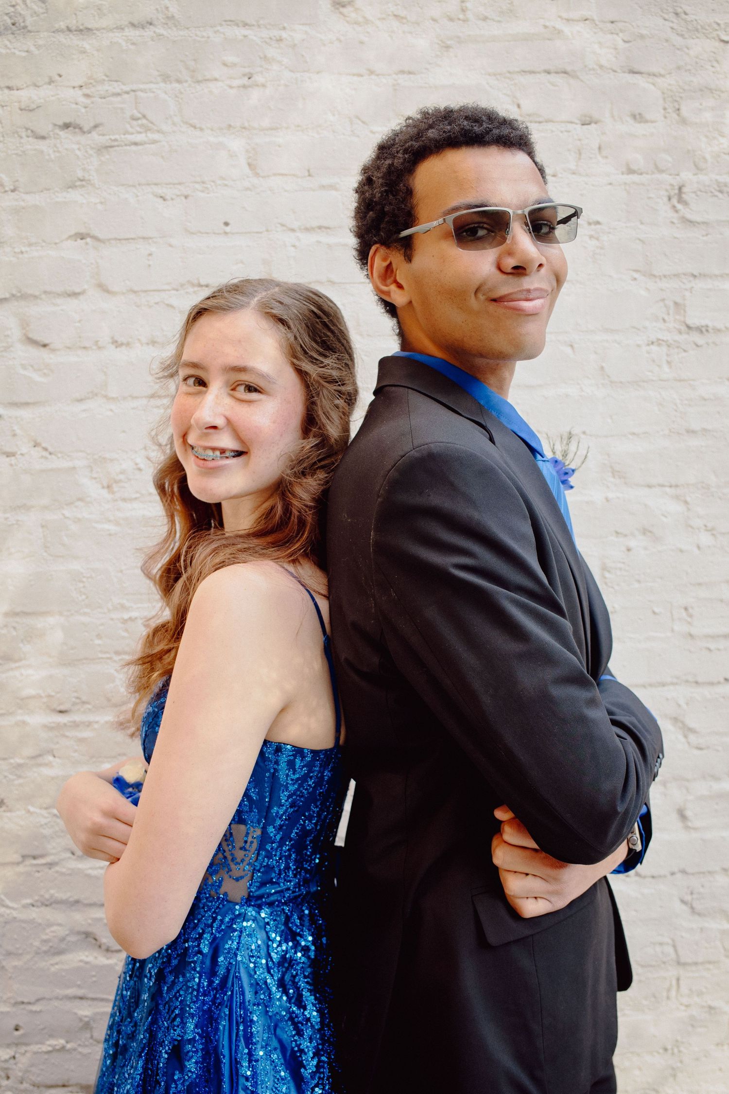 Teen girl in blue sequin dress and boy in black suit with sunglasses pose back-to-back at prom.