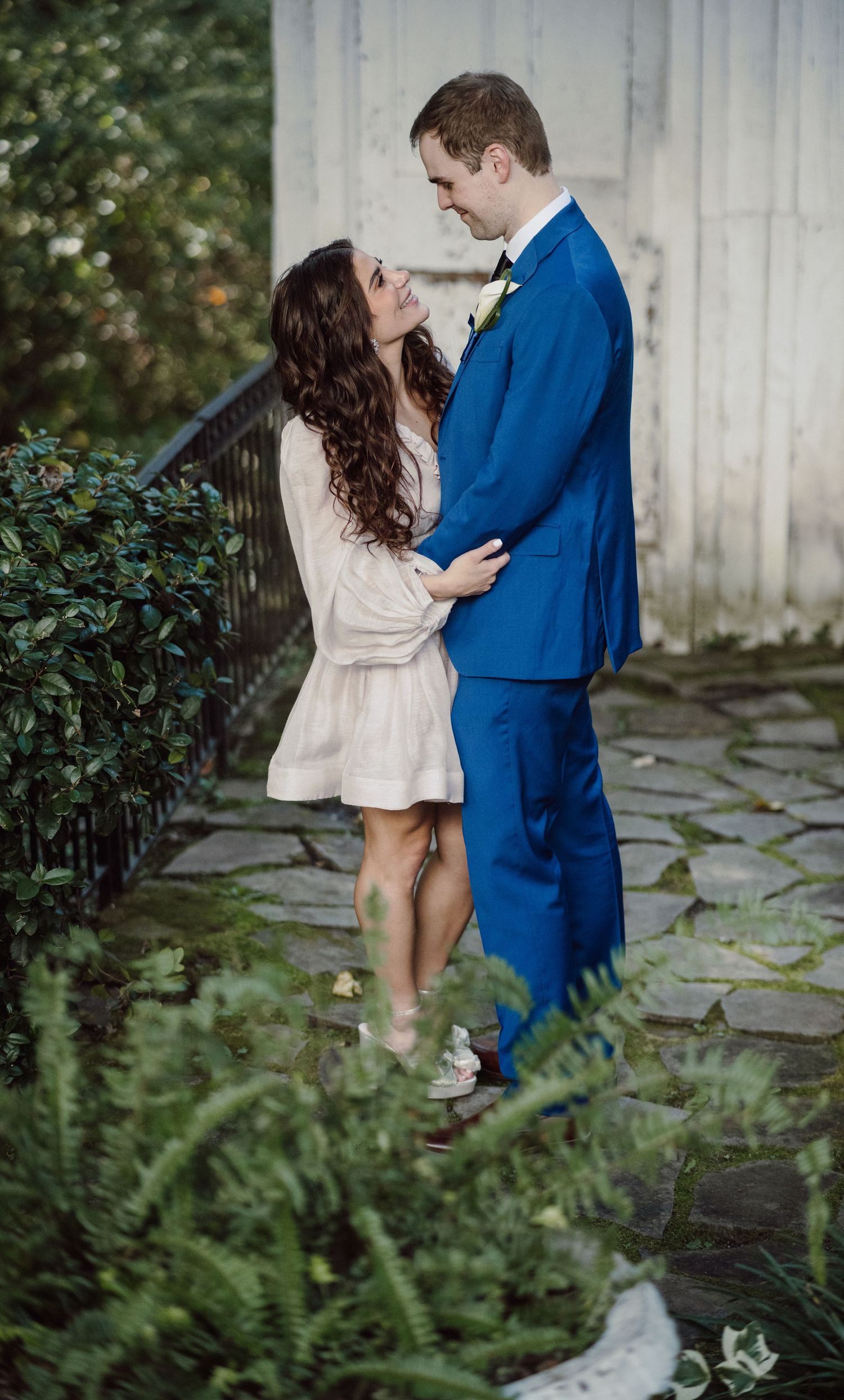Romantic garden photoshoot featuring a couple in blue suit and beige dress among lush ferns and stone pathways.