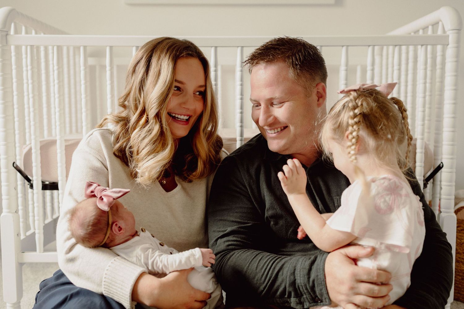 Happy family with newborn baby and toddler girl smiling together in a white nursery setting.