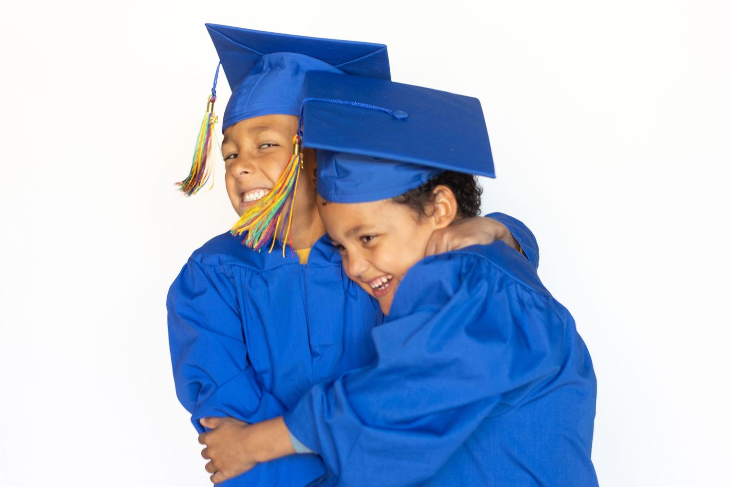 Two young children in blue graduation caps and gowns hugging and smiling against a white background.