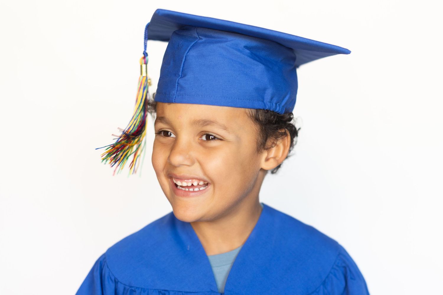 Smiling young child wearing a blue graduation cap and gown, celebrating academic achievement against white background.