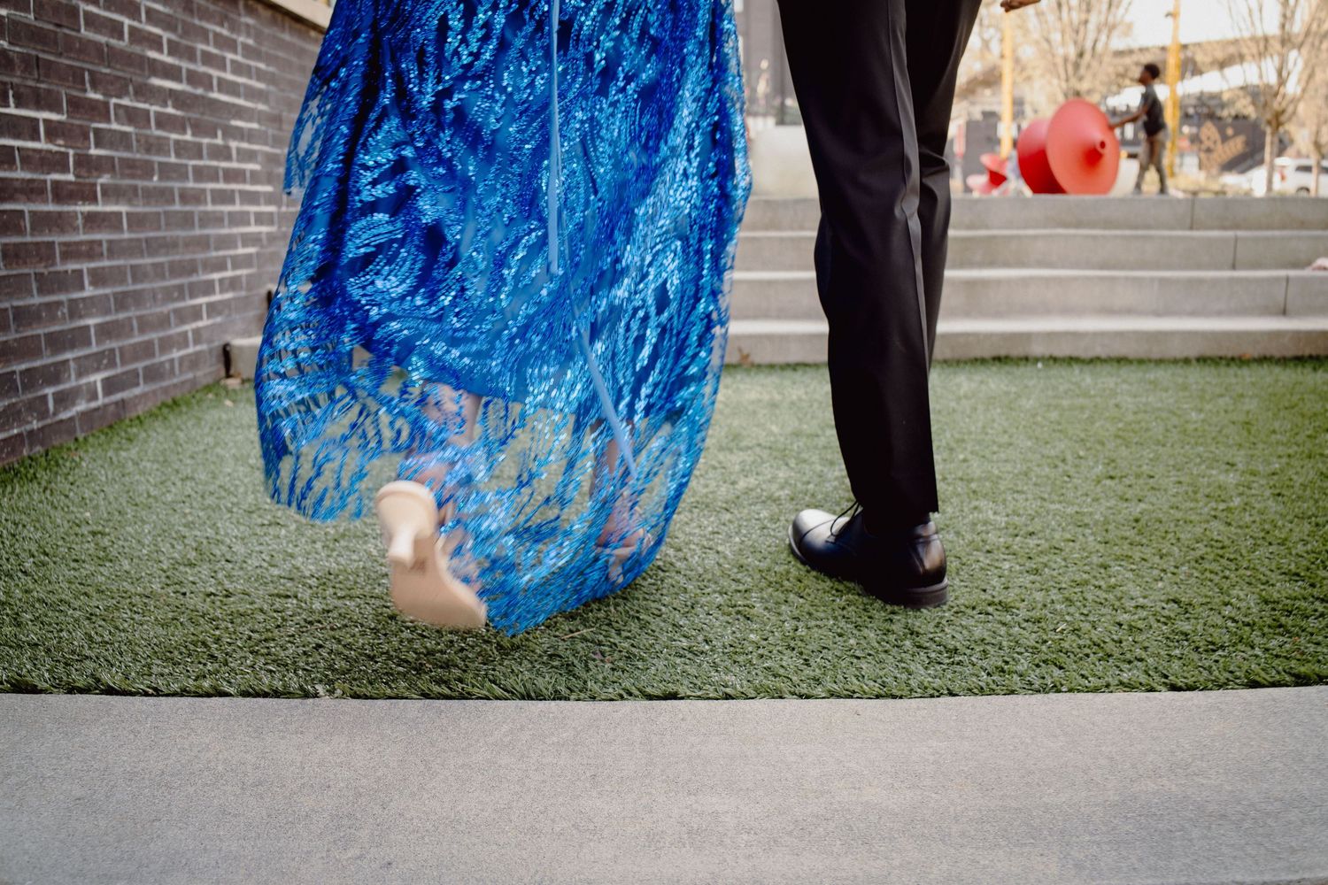 Woman in blue sequin gown and man in black tuxedo walking on green turf, woman holding red balloon.