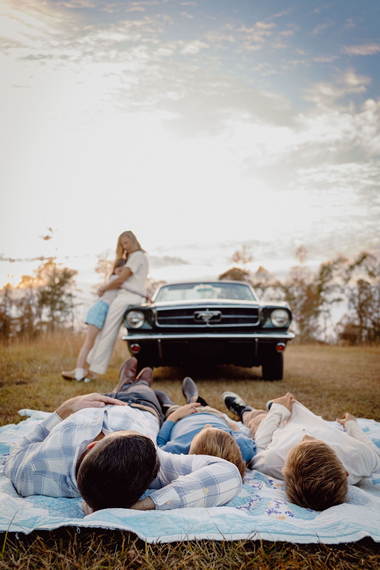 Group of people lying on blankets in a field at sunset with a classic car silhouetted in the background.