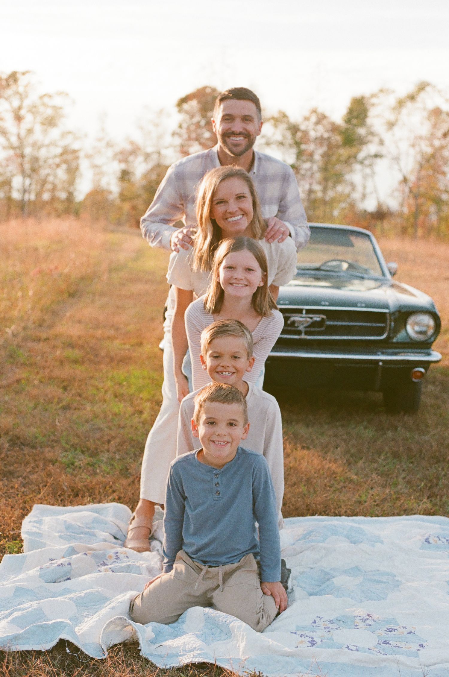 A family sits together on a blanket outdoors during golden hour with a vintage black car visible in the background.