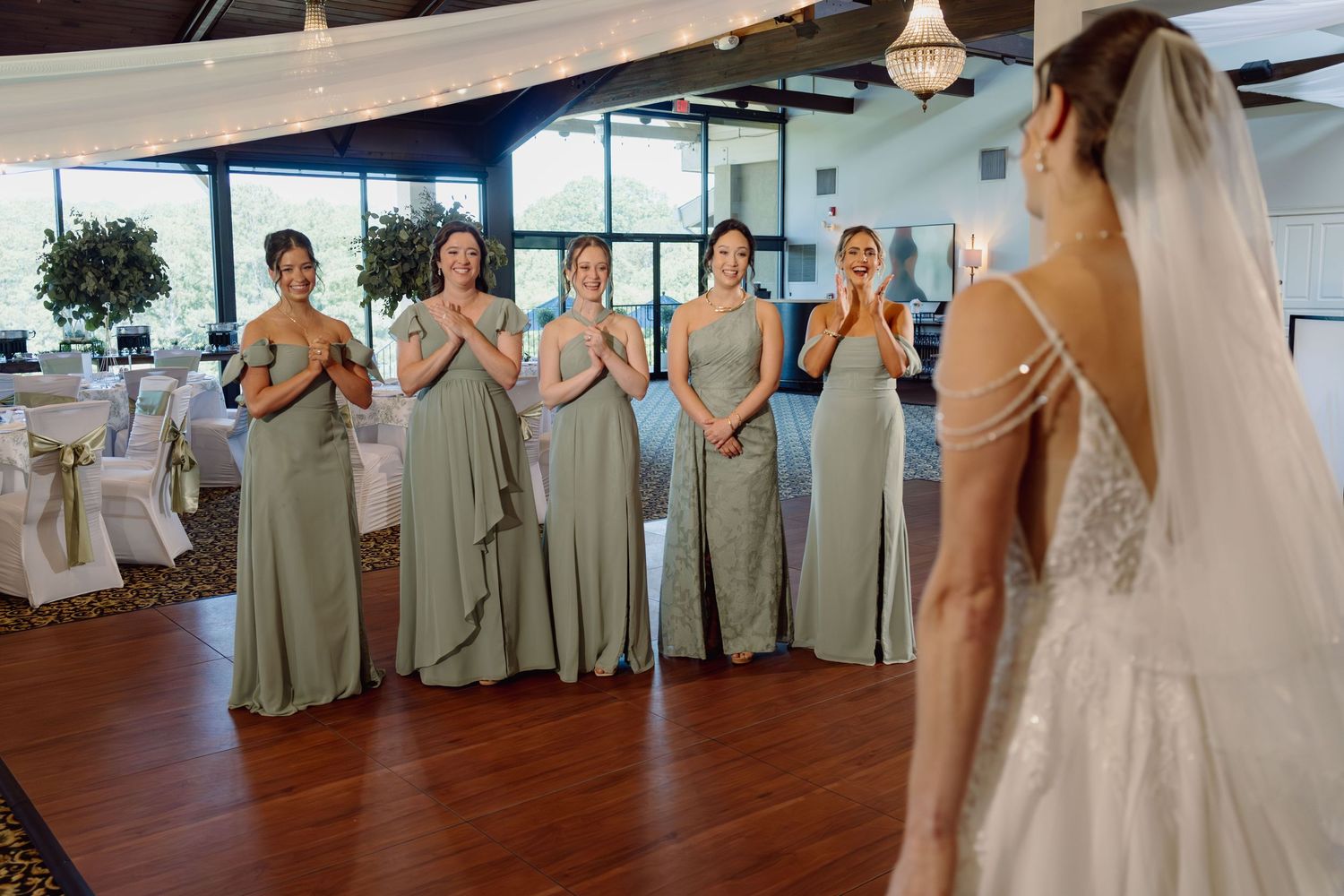 Bride in lace gown reveals herself to emotional bridesmaids in sage green dresses at elegant wedding venue.