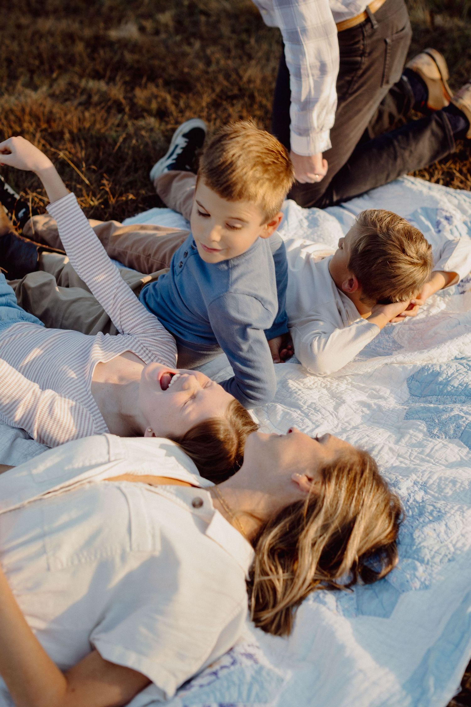 Family relaxing on white blanket outdoors, two women lying down while children play around them in warm sunlight.