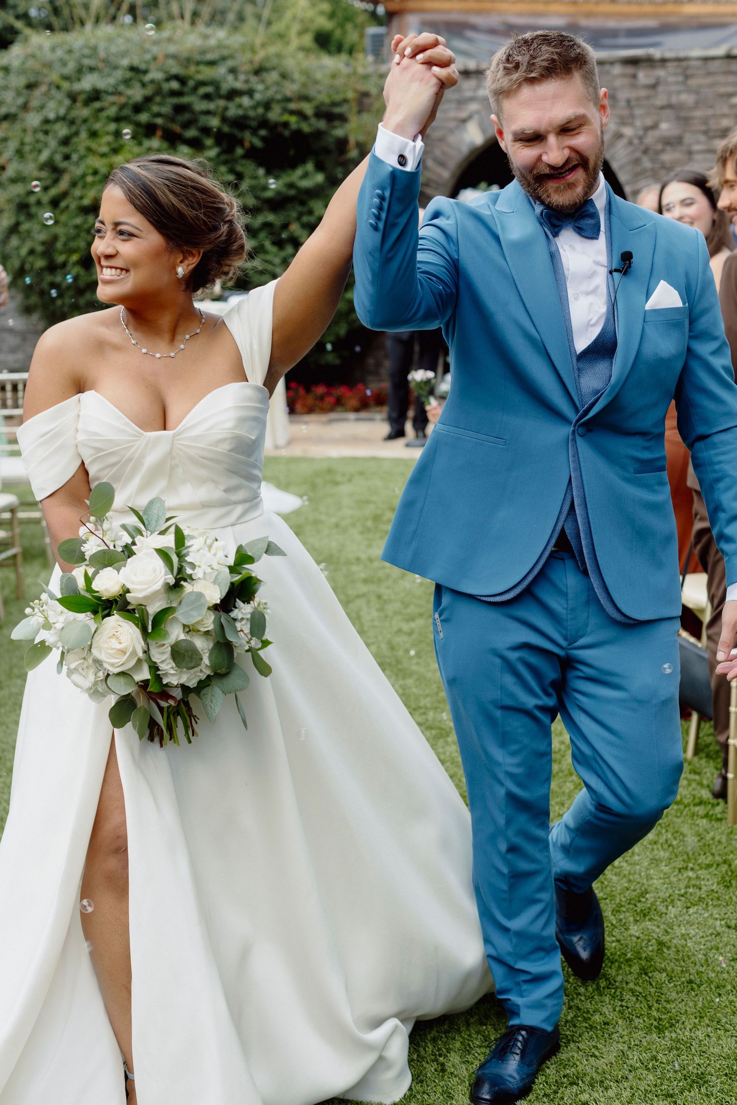 Wedding couple walking on grass in blue suit and white dress with bouquet.
