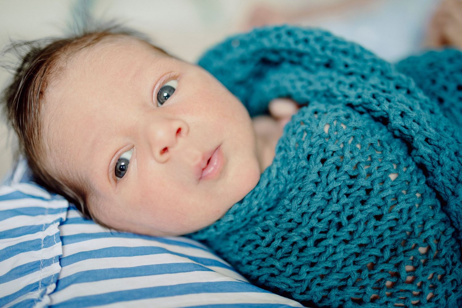 A baby wrapped in a teal knitted blanket lies on striped bedding looking upward with bright eyes.