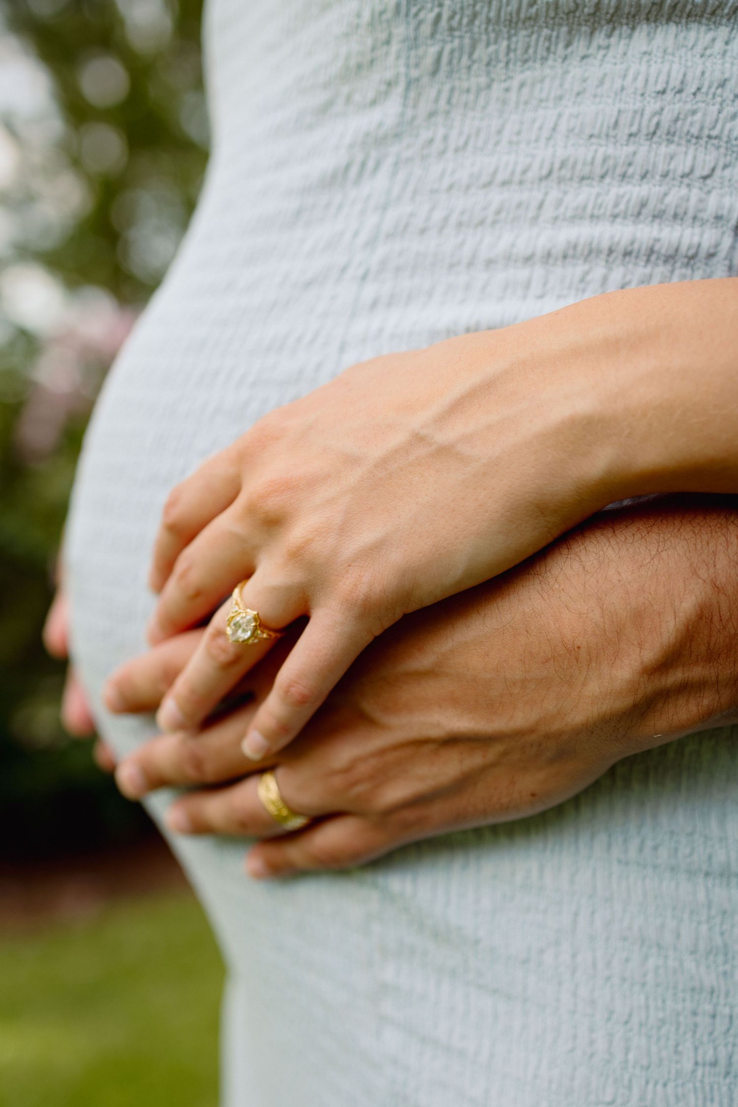 Pregnant woman in light blue dress with hands clasped over baby bump, wearing gold engagement and wedding rings.