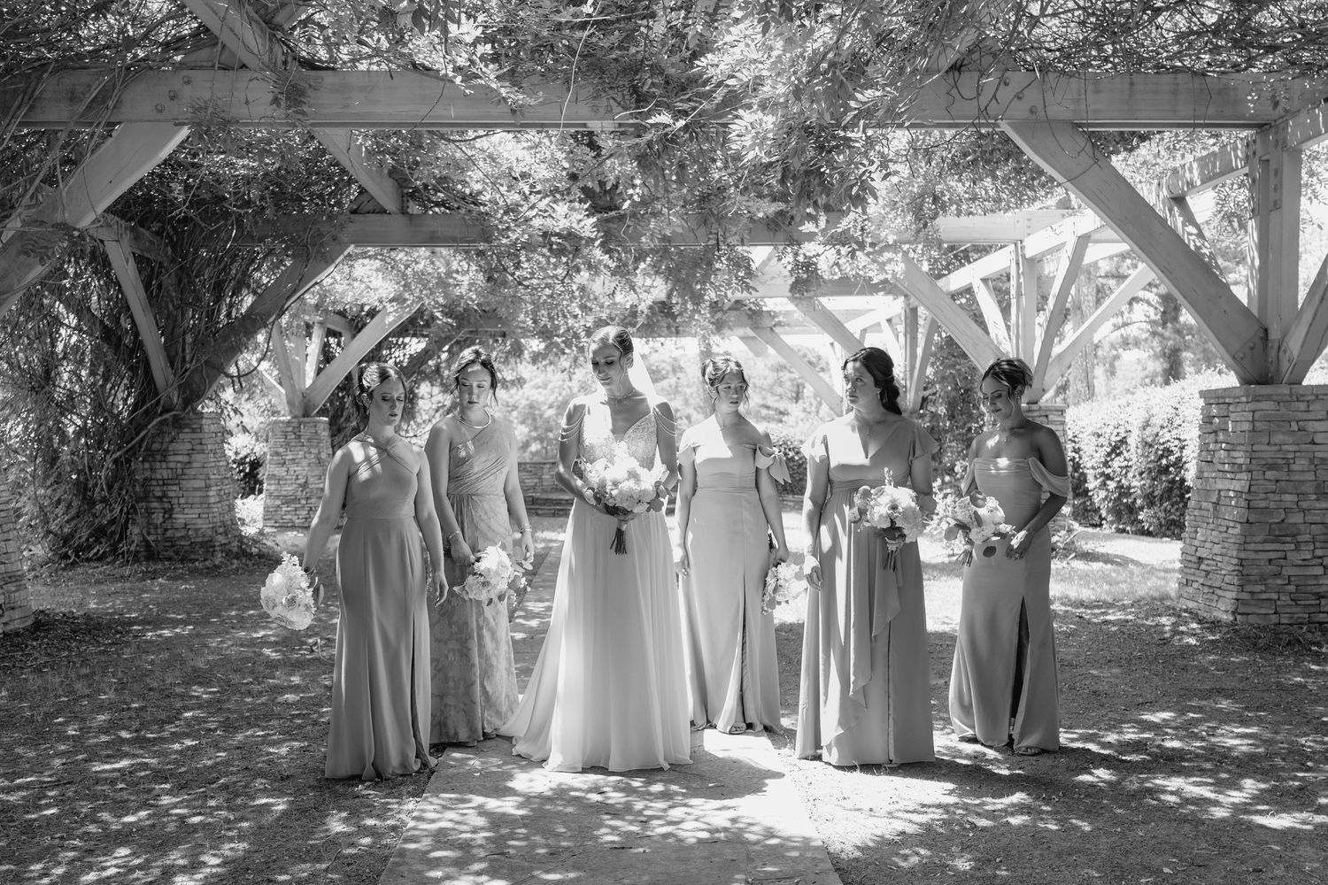 Black and white photo of a bride and bridesmaids in long gowns holding bouquets under a wooden pergola outdoors.