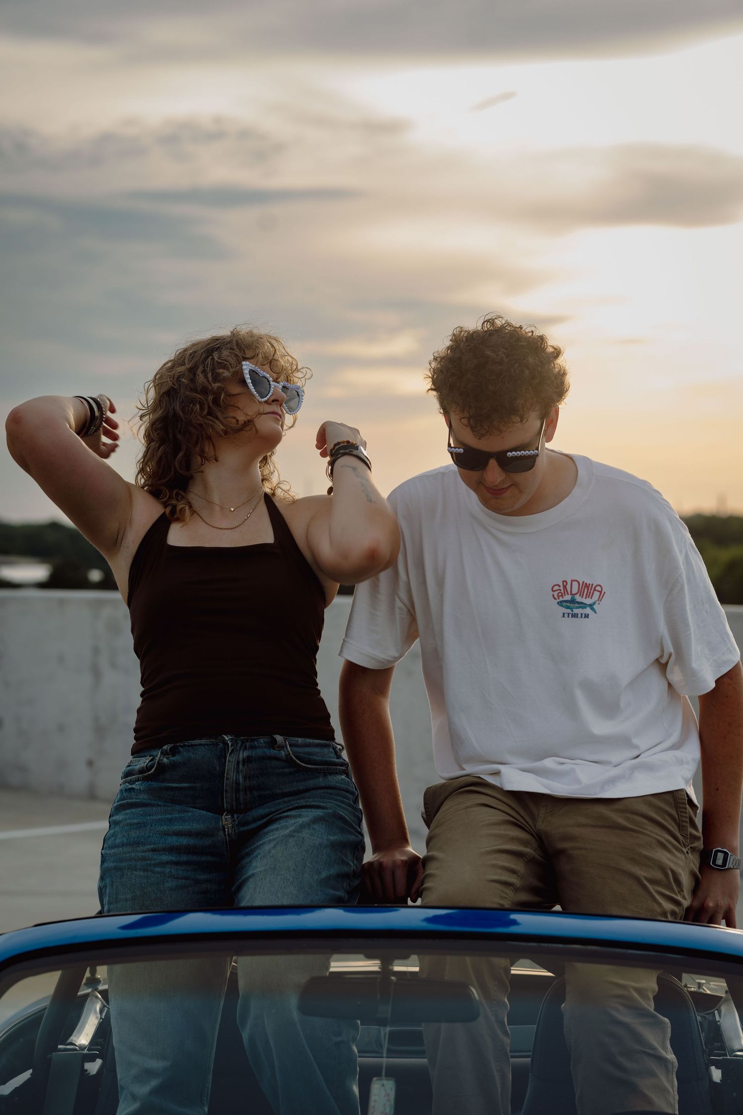 Two young friends pose on a blue convertible car at sunset, wearing sunglasses in casual summer outfits.