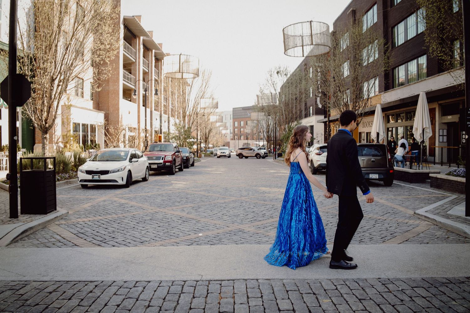 Couple in formal wear walking on a cobblestone street, woman in blue gown, urban backdrop.