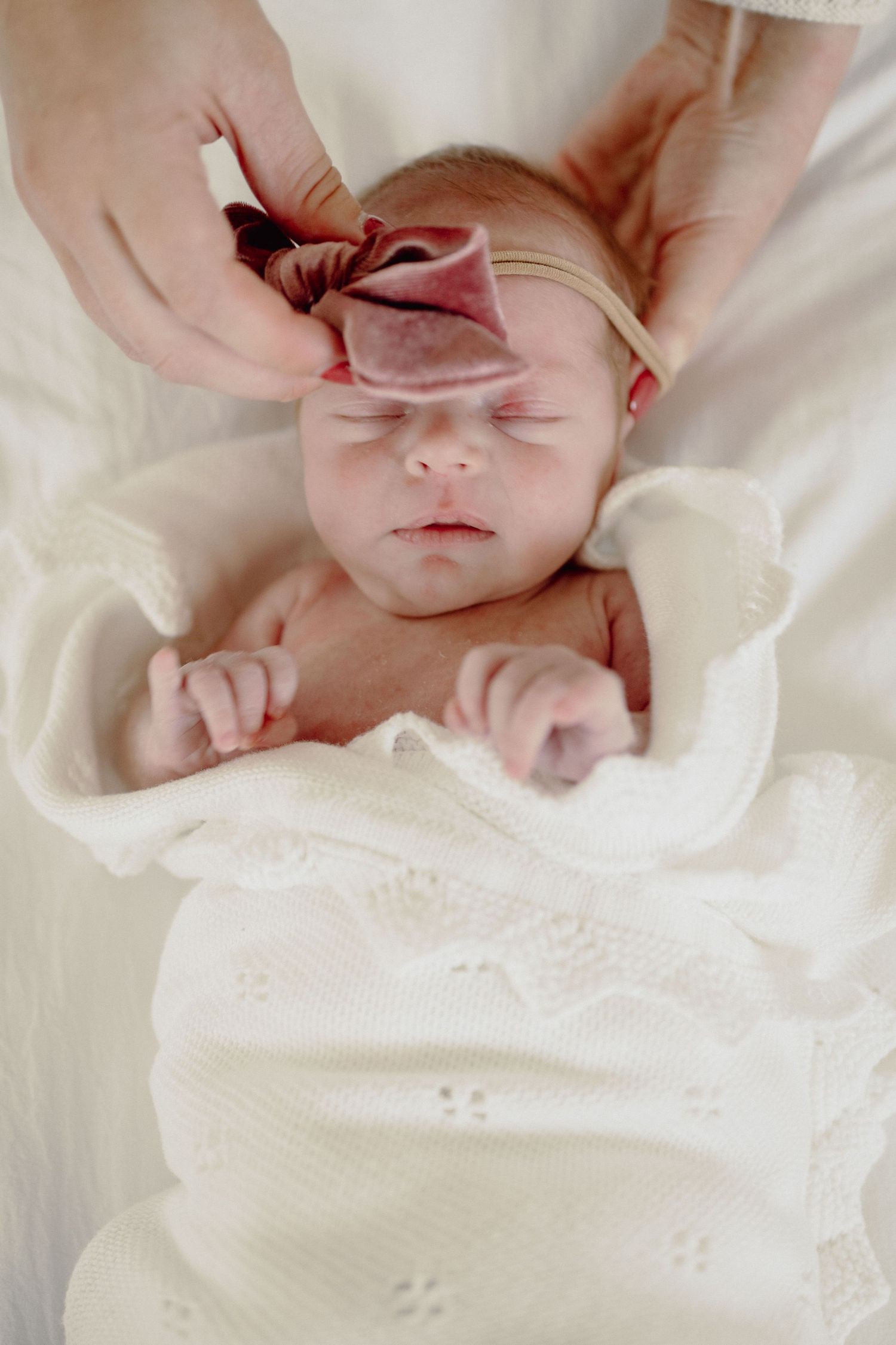Newborn baby in white knit blanket with pink velvet bow headband being placed by mother's hands.