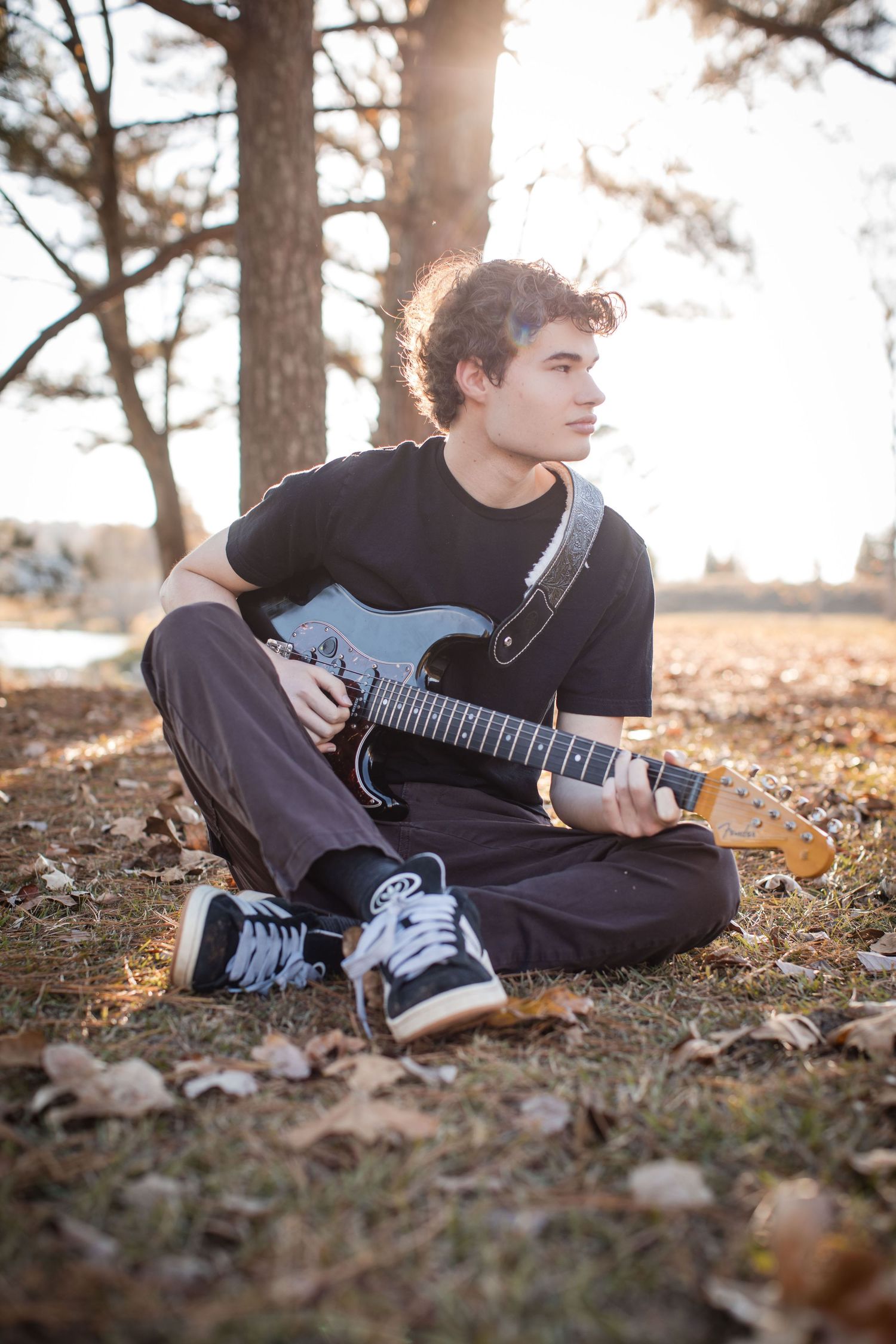 Young man sitting cross-legged outdoors playing electric guitar among autumn leaves with sunlight filtering through trees.