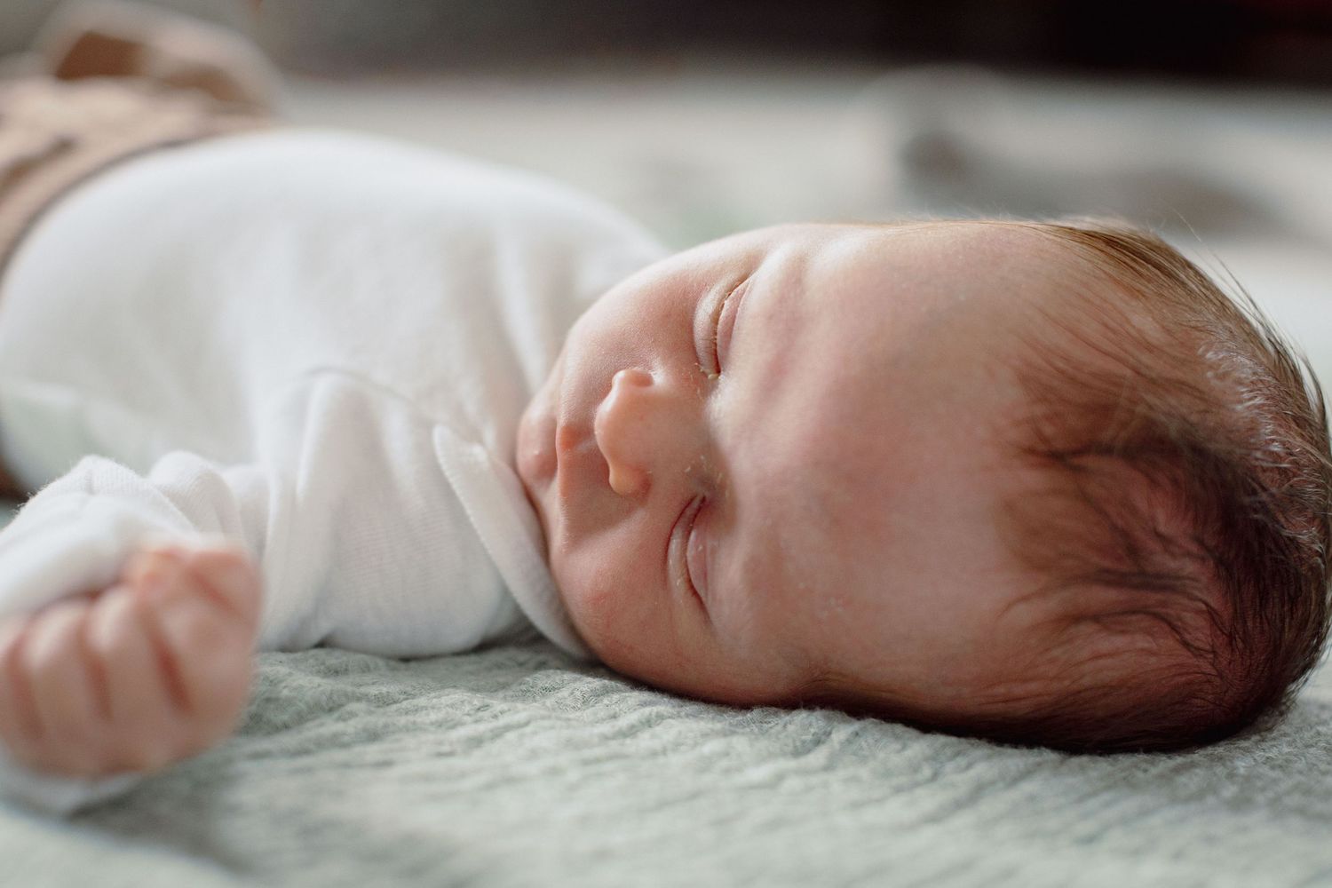 Peaceful newborn baby sleeping on a soft gray blanket, wearing a white onesie with tiny fists curled up.