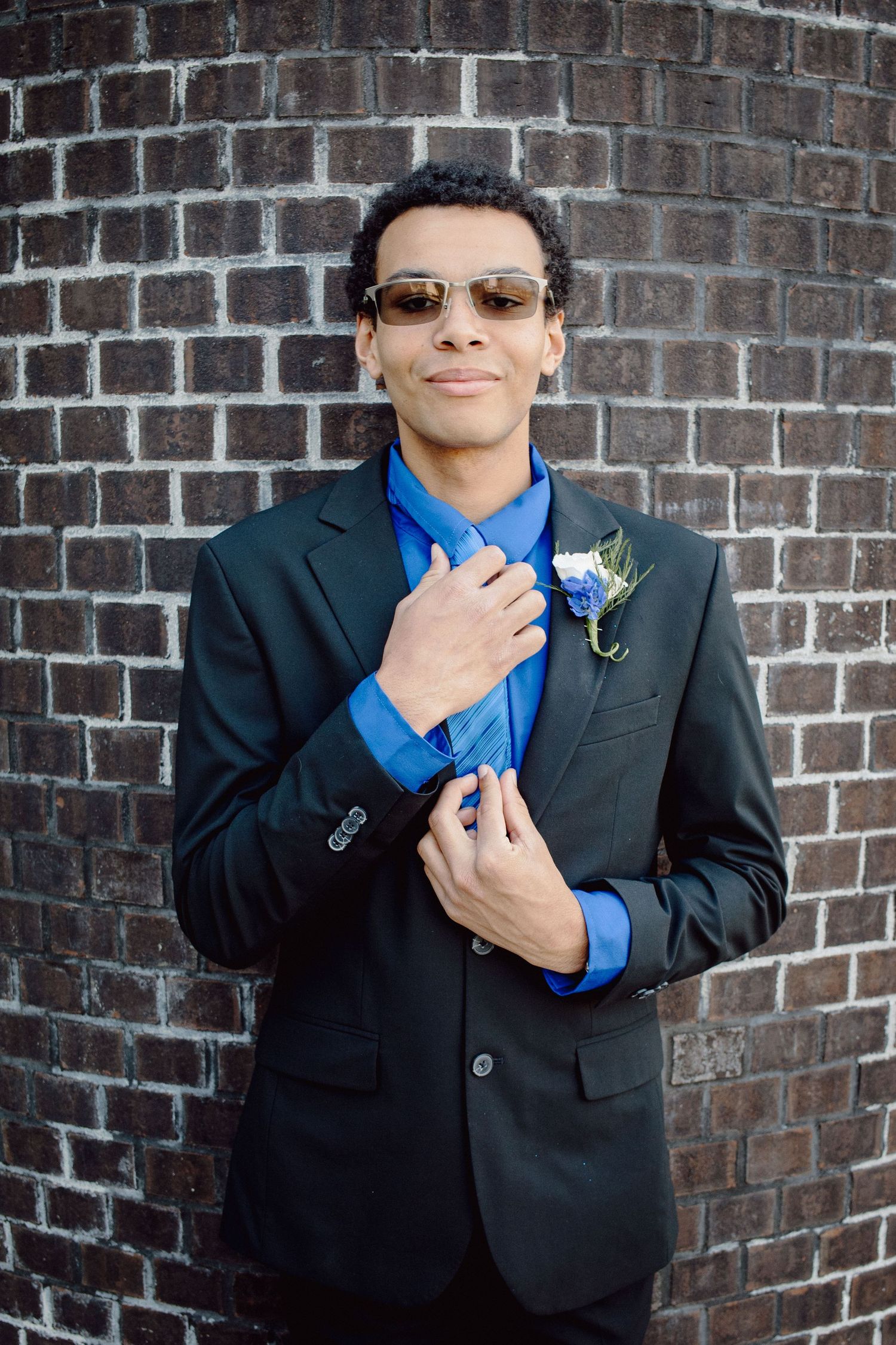A stylish young man in a black suit, blue shirt, and sunglasses poses with a boutonniere against a brick wall.