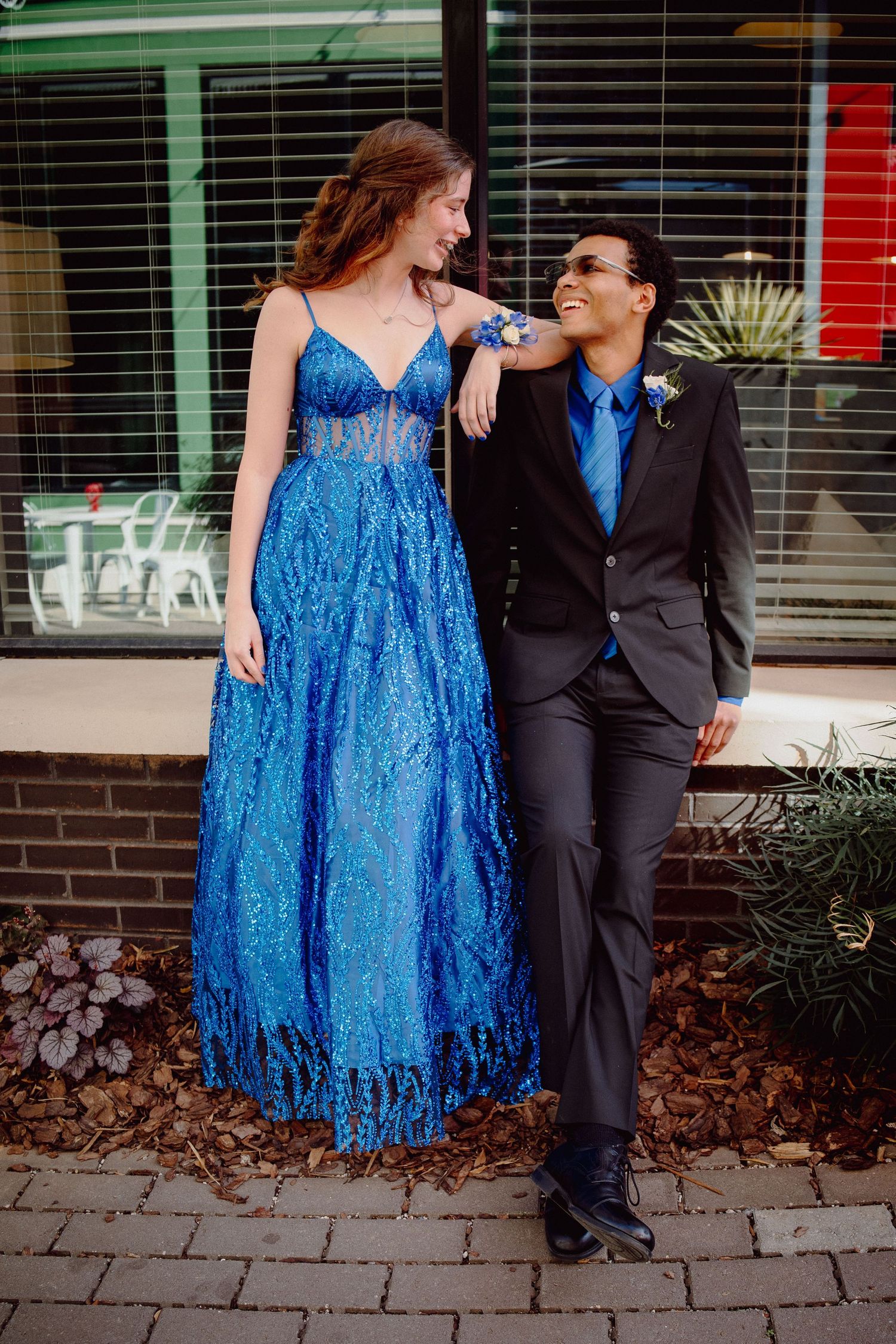 A couple in matching blue prom attire pose together outside, she in a sequined gown and he in a suit with boutonniere.