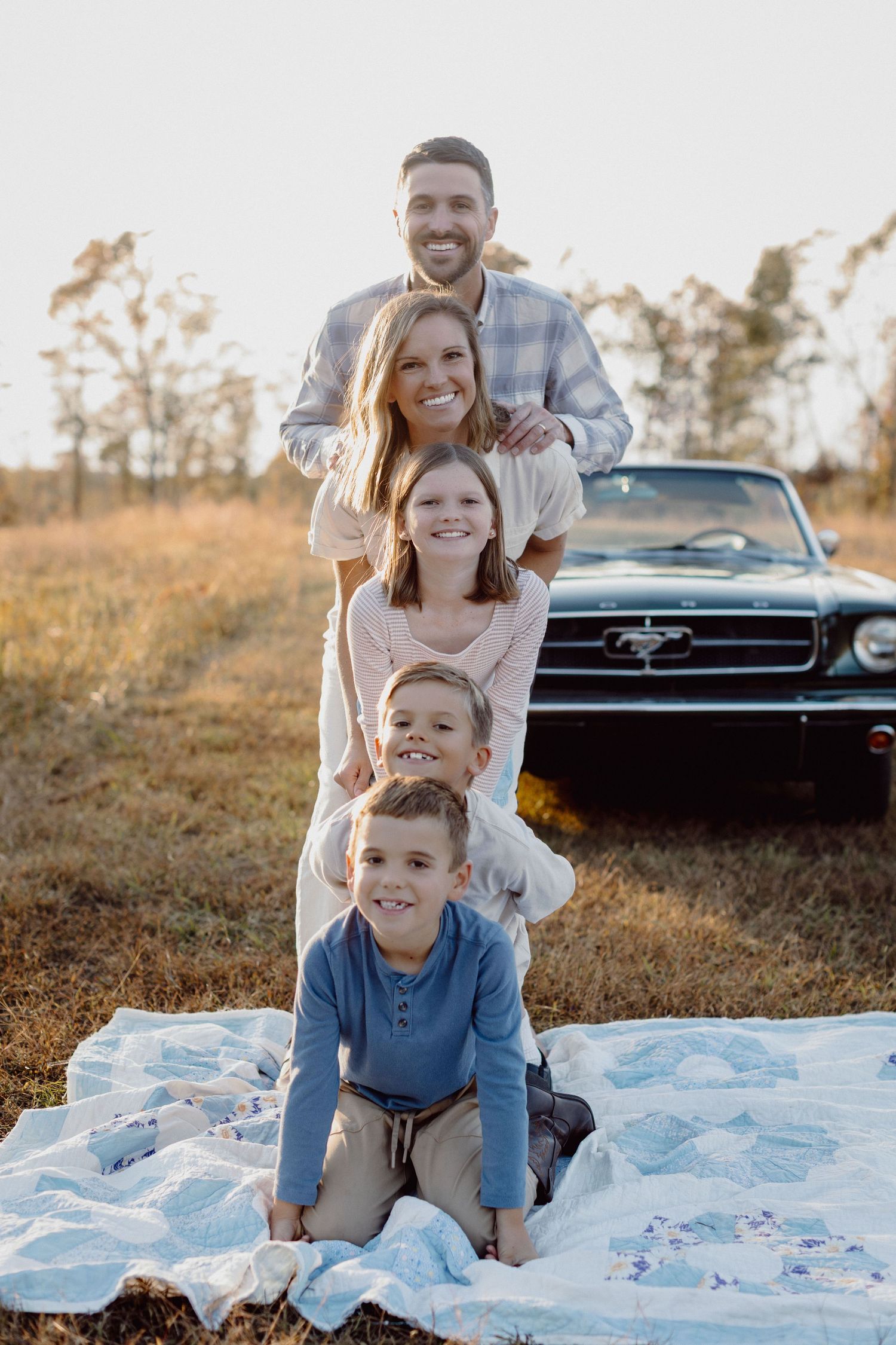 Family sits together on blanket in field at sunset with vintage black Chevrolet car in background.
