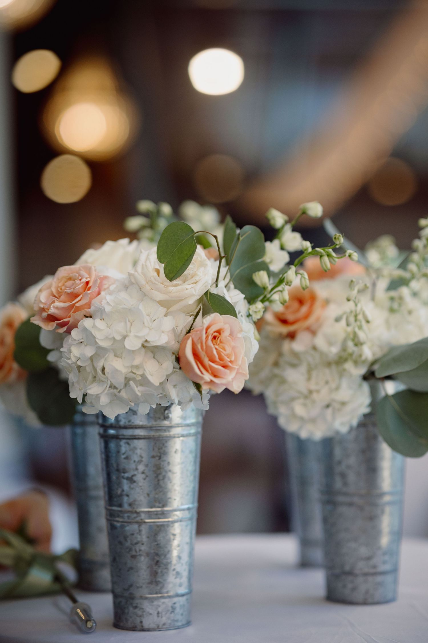 Galvanized metal vases with white hydrangeas and peach roses centerpieces on a white table.