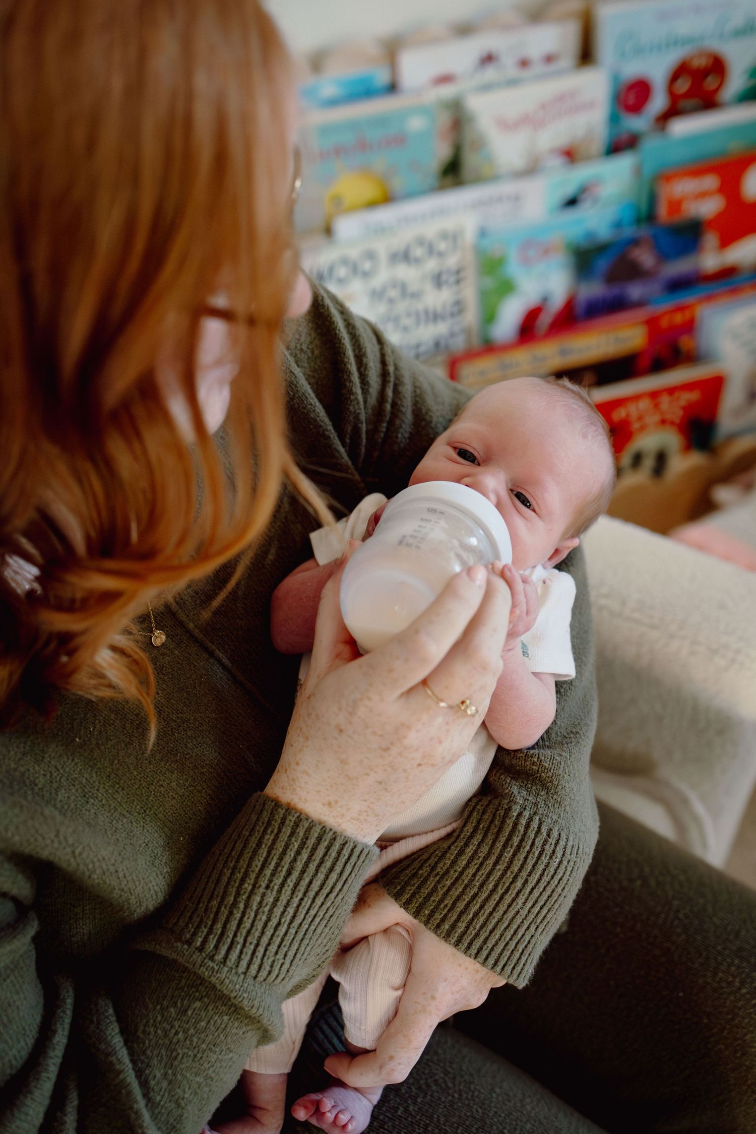 A small pink plush toy being held in hands wearing a green knit sweater with children's books in the background.