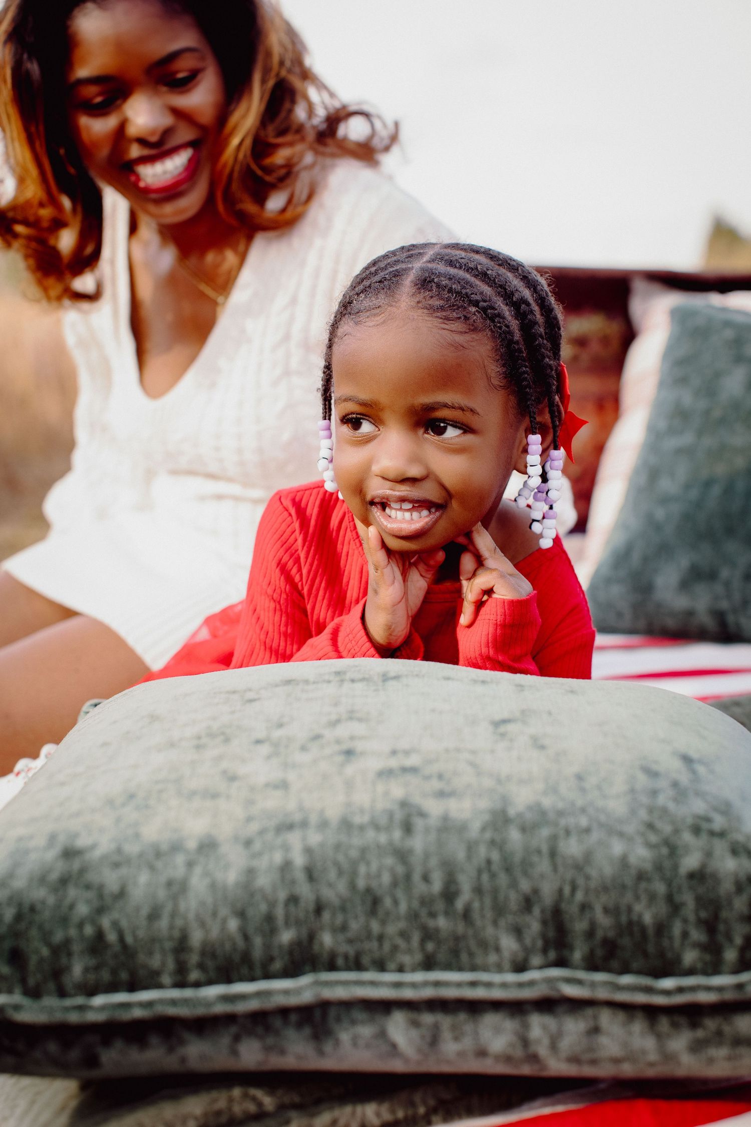 A young child in a red sweater smiles playfully while resting their head on a gray couch cushion.