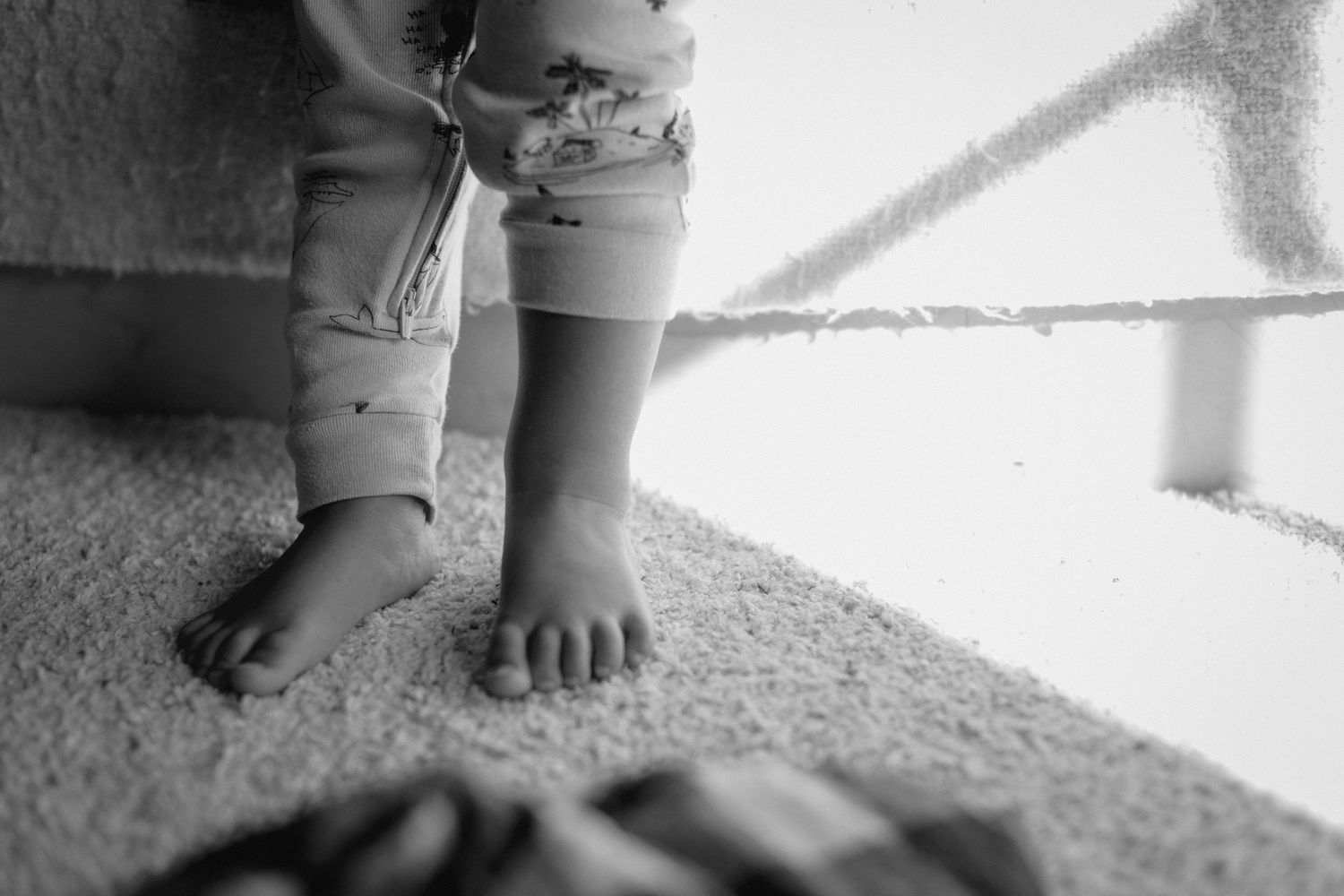 Black and white photo of a baby standing on carpet near a crib, showing tiny bare feet and chubby legs.