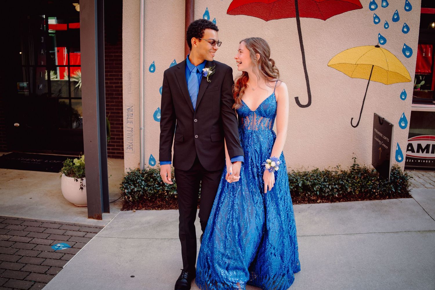 A couple in prom attire poses on a sidewalk, the woman in a blue gown and the man in a dark suit with blue tie.