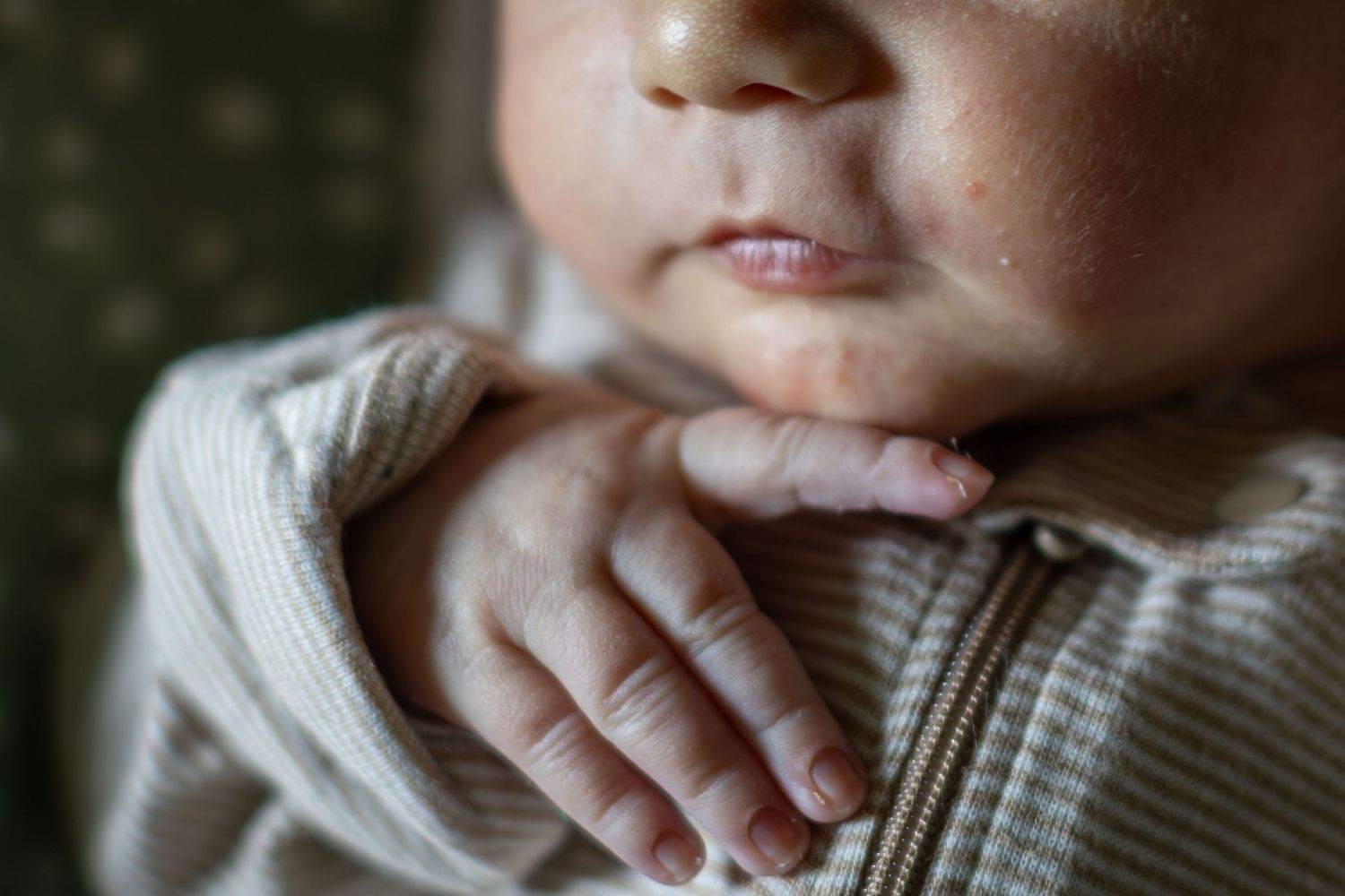 Close-up of a newborn baby's tiny hands and chubby chin wearing a beige ribbed zip-up onesie.