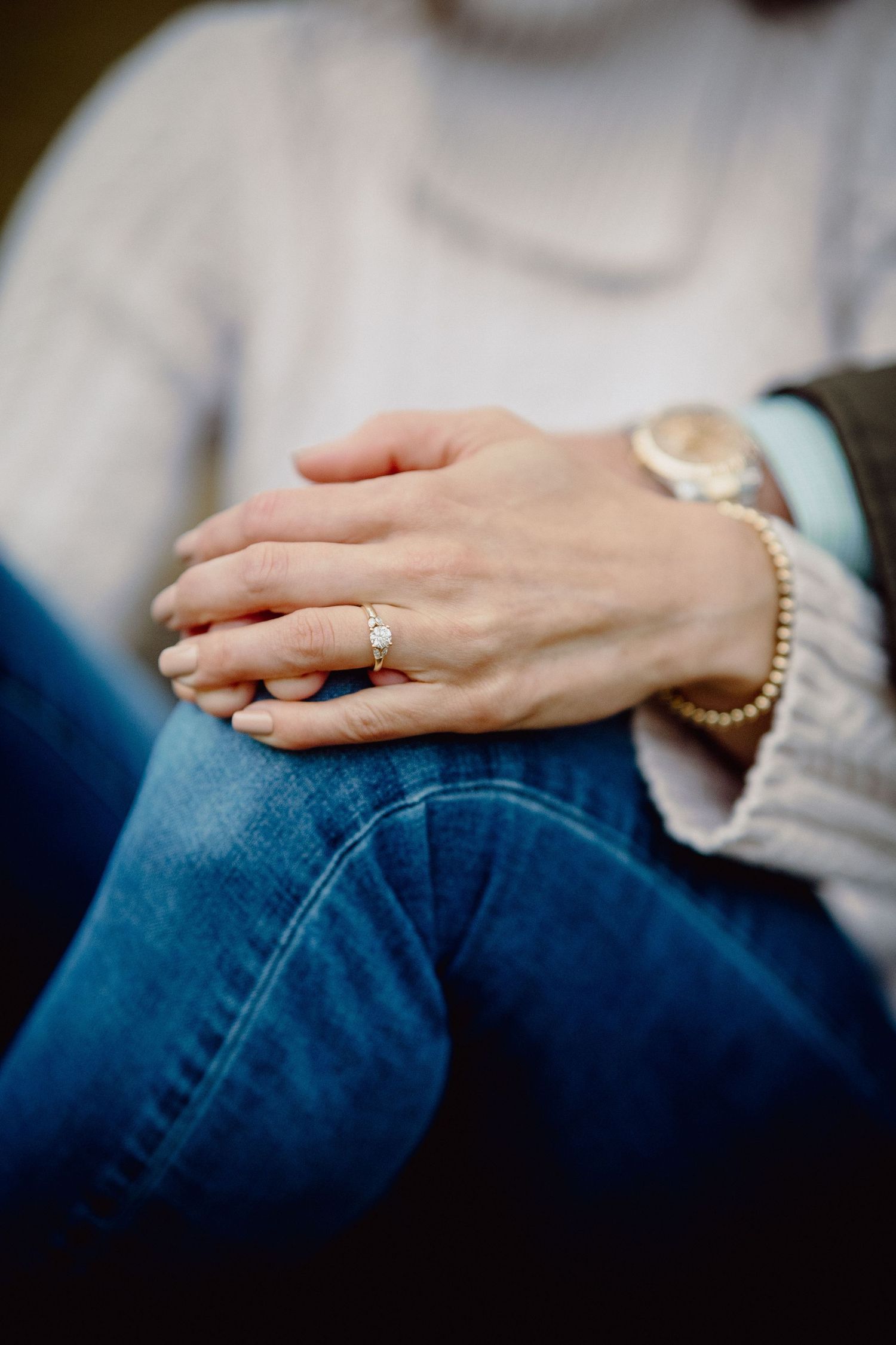 A hand wearing a delicate ring and bracelet rests gently on blue denim jeans.