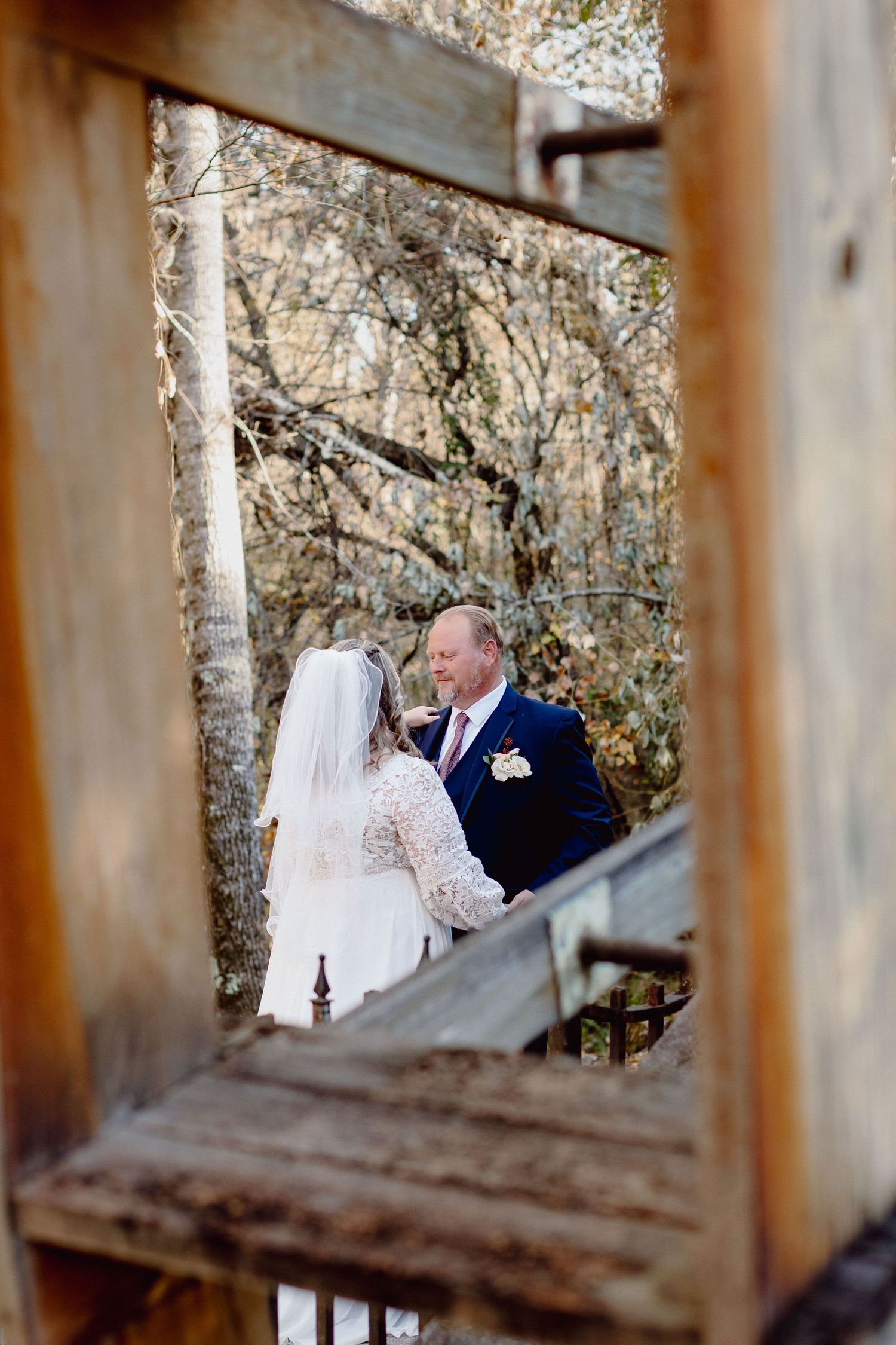 A romantic wedding moment captured through wooden railings with a rustic stone wall backdrop.