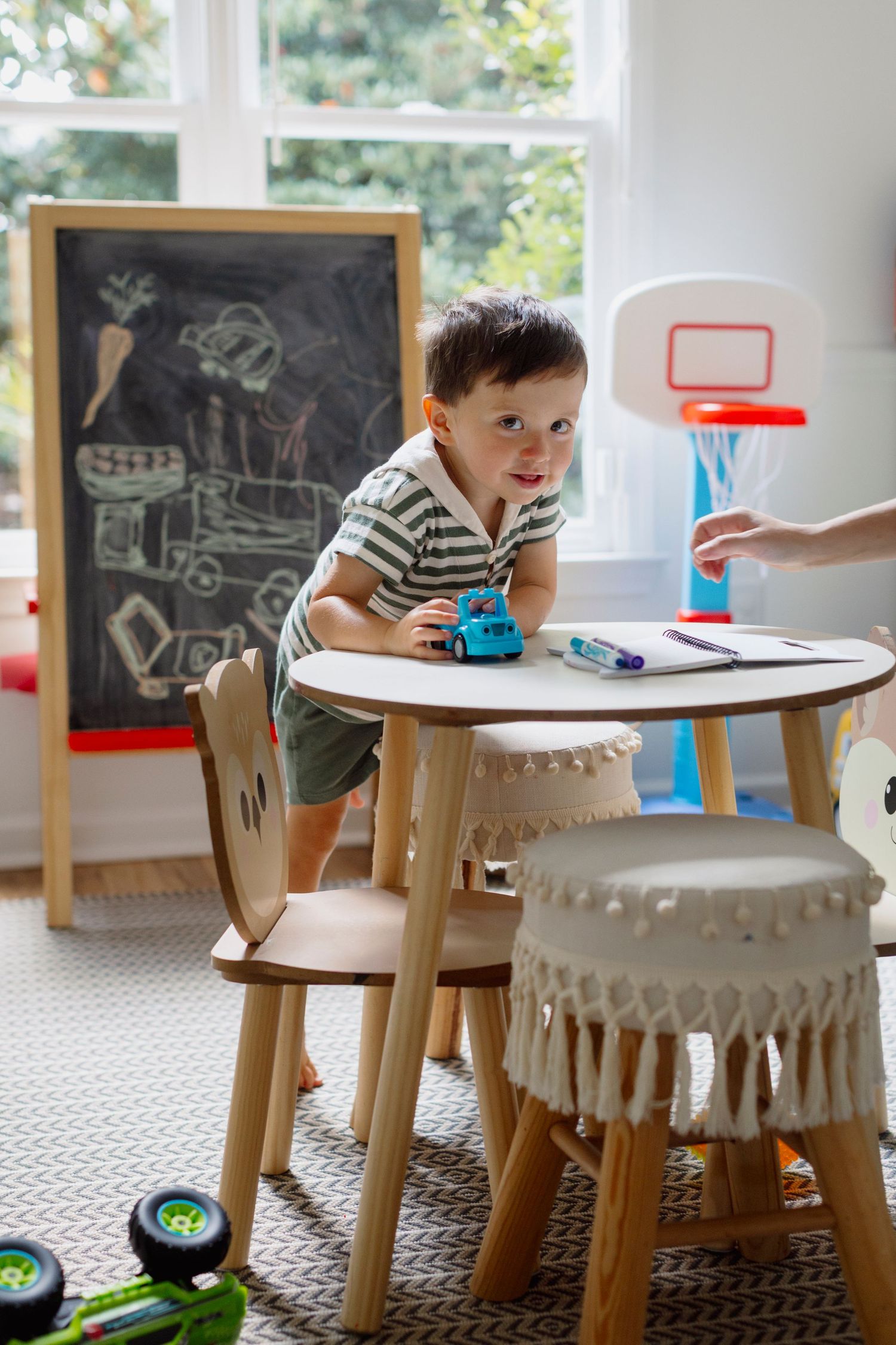 Child plays at wooden table in playroom with chalkboard and basketball hoop visible in background.