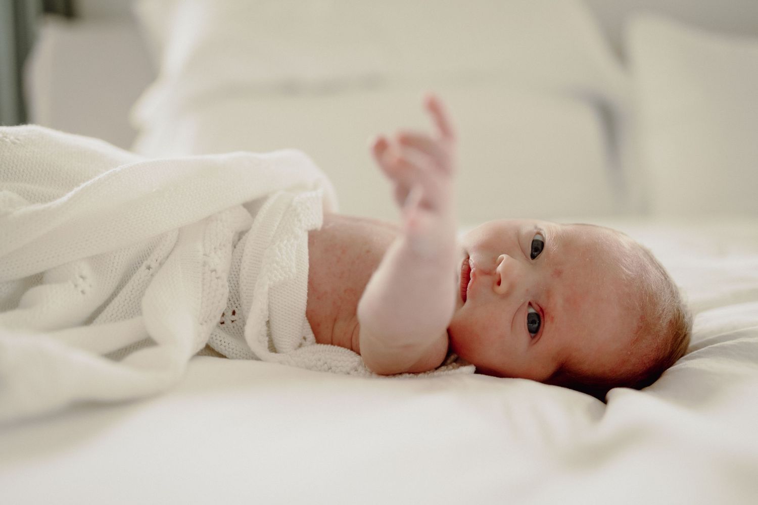 A baby lying on white bedding stretches their tiny arm while wrapped in a soft blanket.