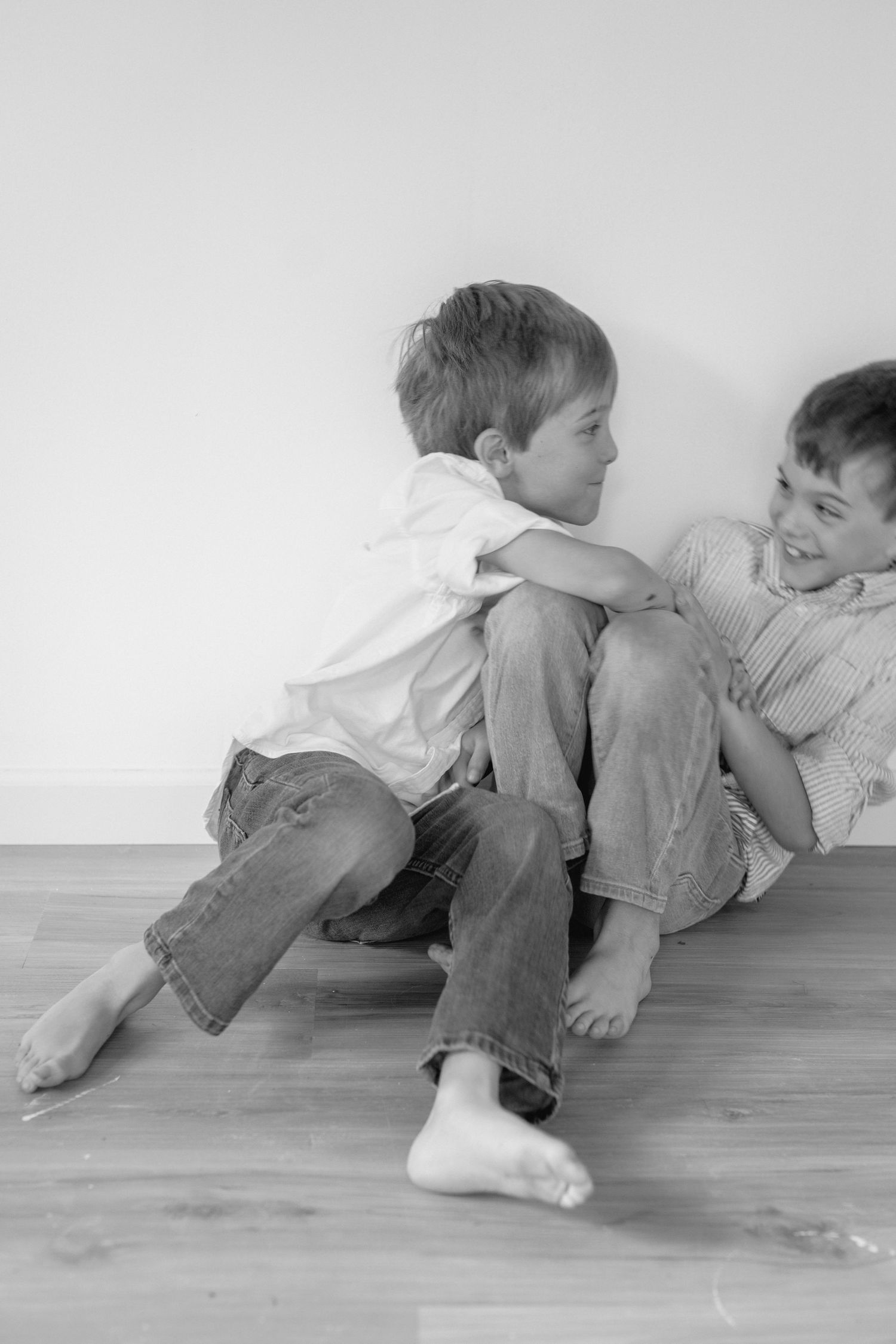 Two children in casual clothing lay side by side on a hardwood floor in a black and white photograph.