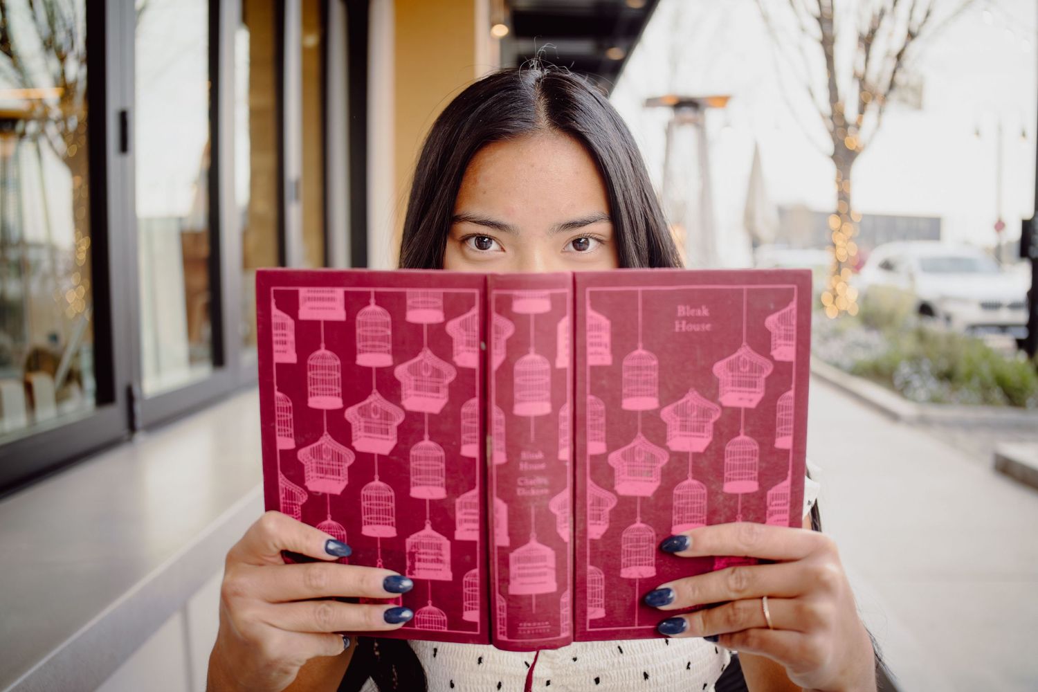 Woman peering over a red decorative book with birdcage pattern, holding it up to cover her face outdoors.