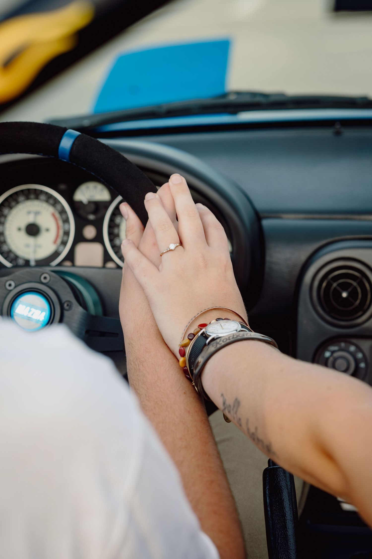 Two hands touching on a car gear shift, one wearing a ring and bracelets, inside a sports car interior.