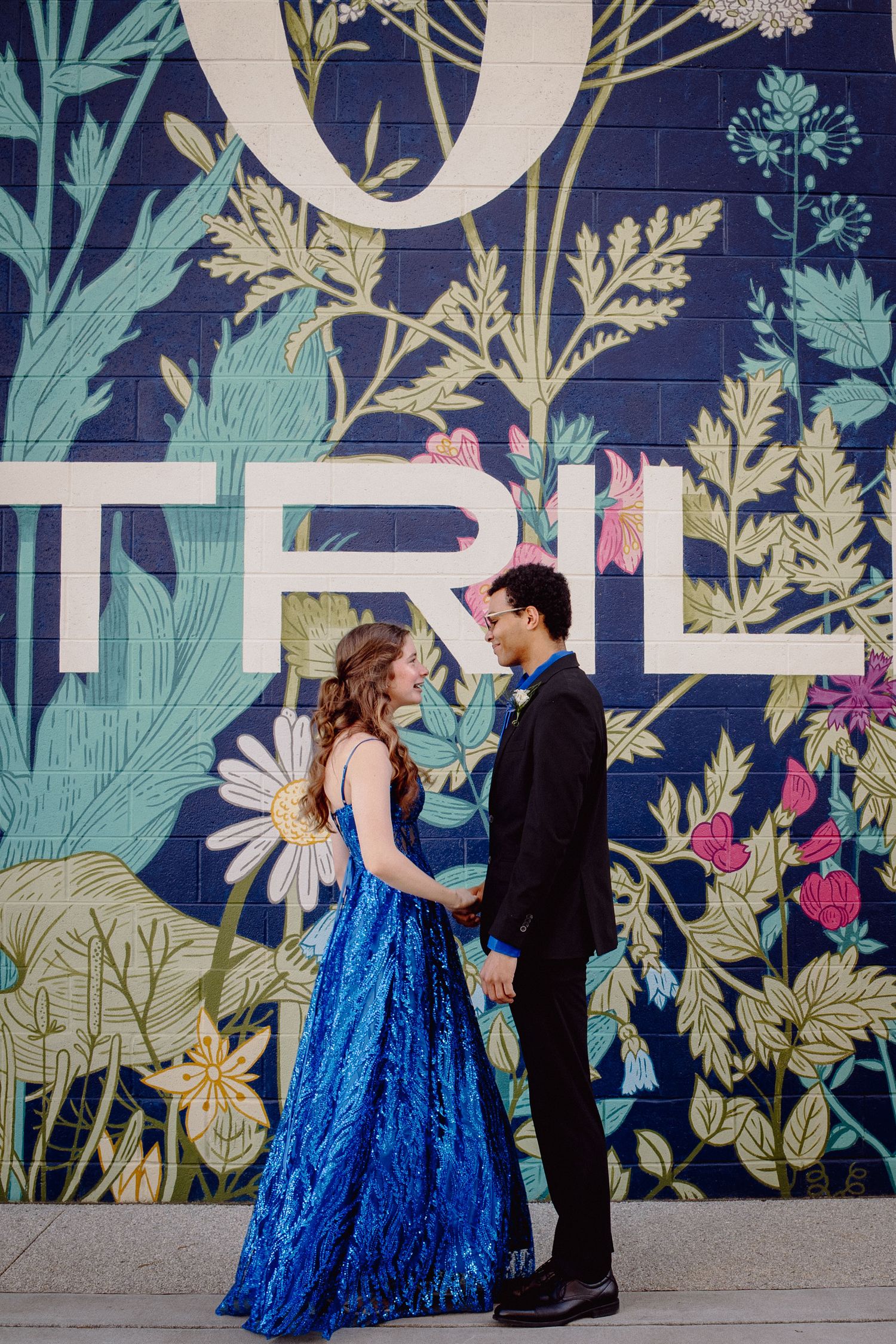 Prom couple holding hands in blue sequin dress and black suit before a navy floral mural with bold lettering.