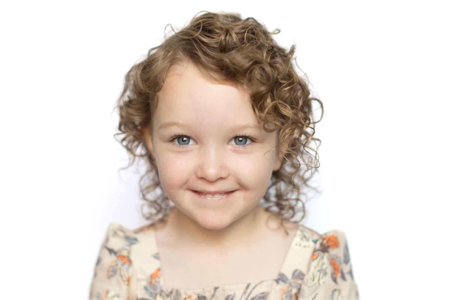 Smiling young girl with curly brown hair wearing a floral dress, posing for a portrait photo on white background.