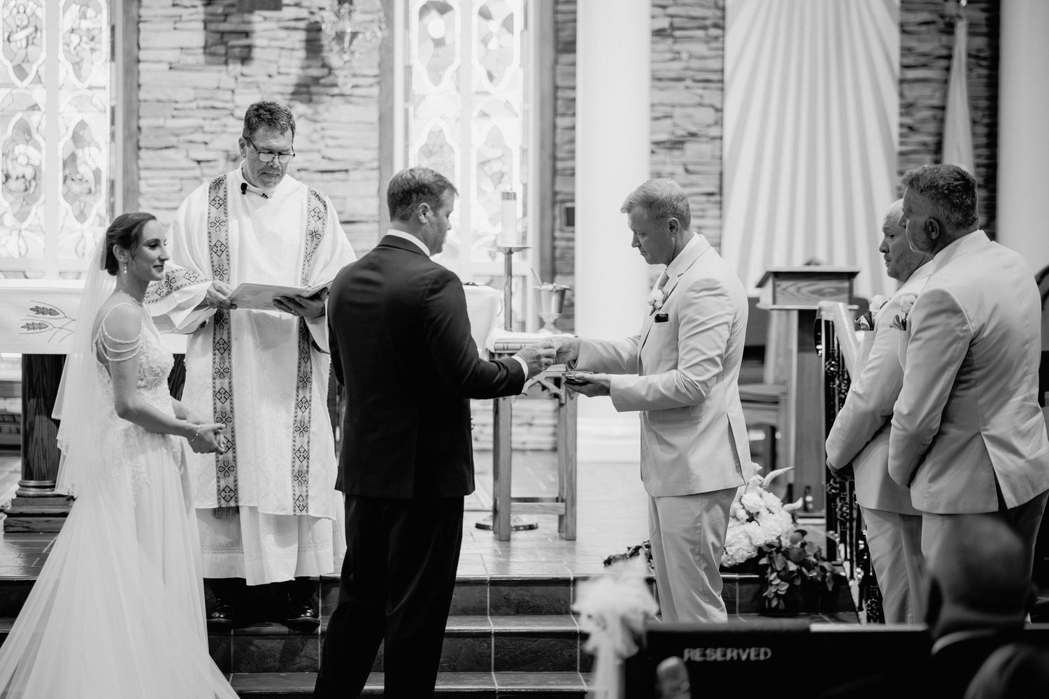 Black and white wedding ceremony photo of bride and groom exchanging rings at church altar with officiant.