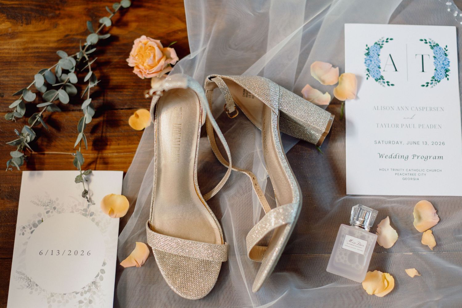 Elegant bridal flat lay with glitter heels, wedding program, perfume, rose petals, and eucalyptus on wood.