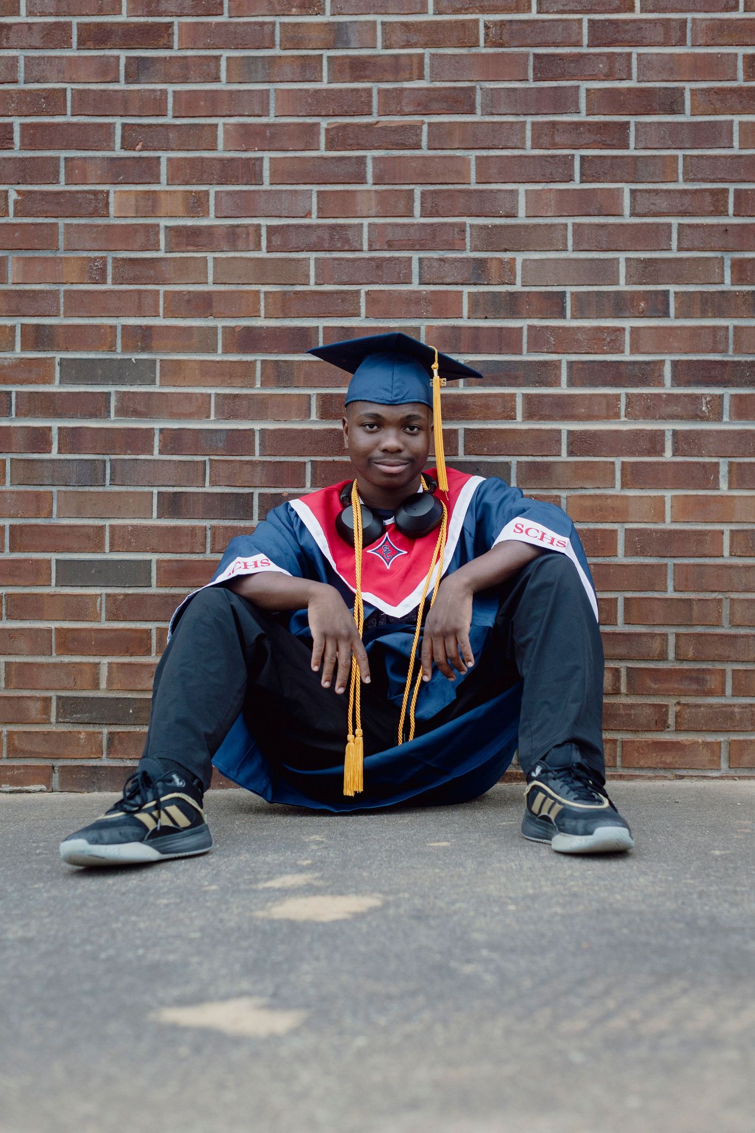 Graduate in cap and gown sitting against brick wall, wearing blue robe with red and gold accents.
