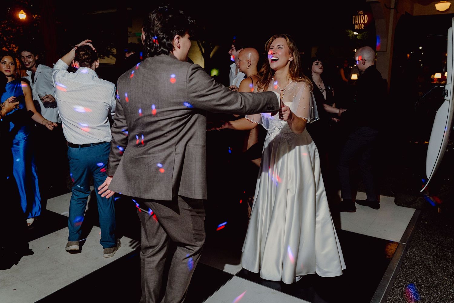 People dancing and celebrating at a dimly lit indoor reception party with purple lighting.
