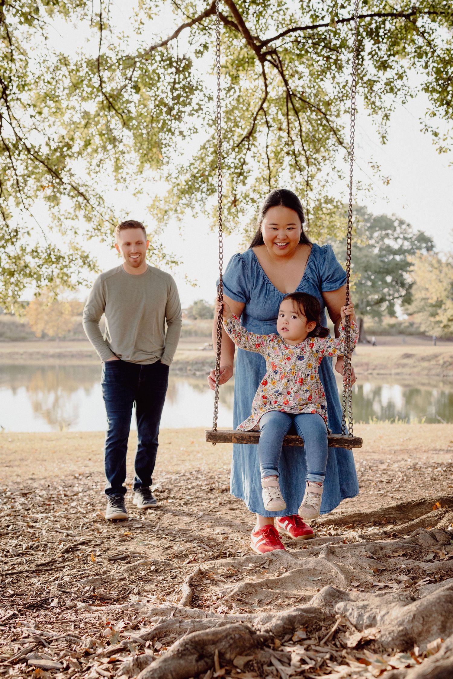 A child enjoys a swing push at a scenic park with tree roots visible in the foreground near a peaceful lake.
